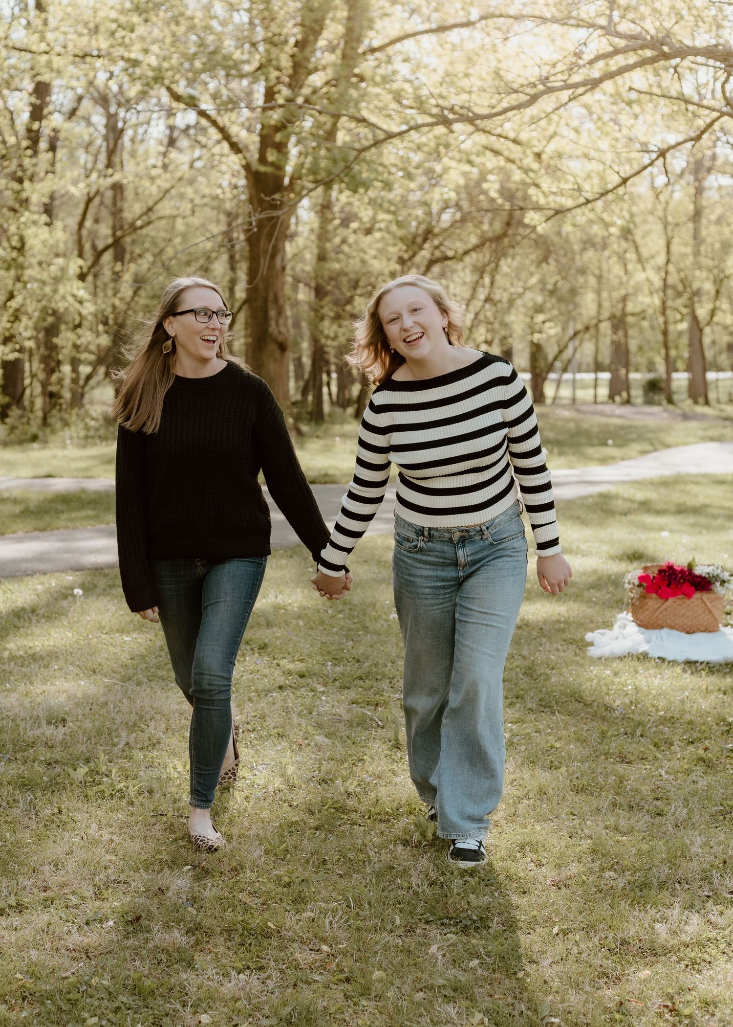 Two young women holding hands and laughing while walking through a sunny green park.