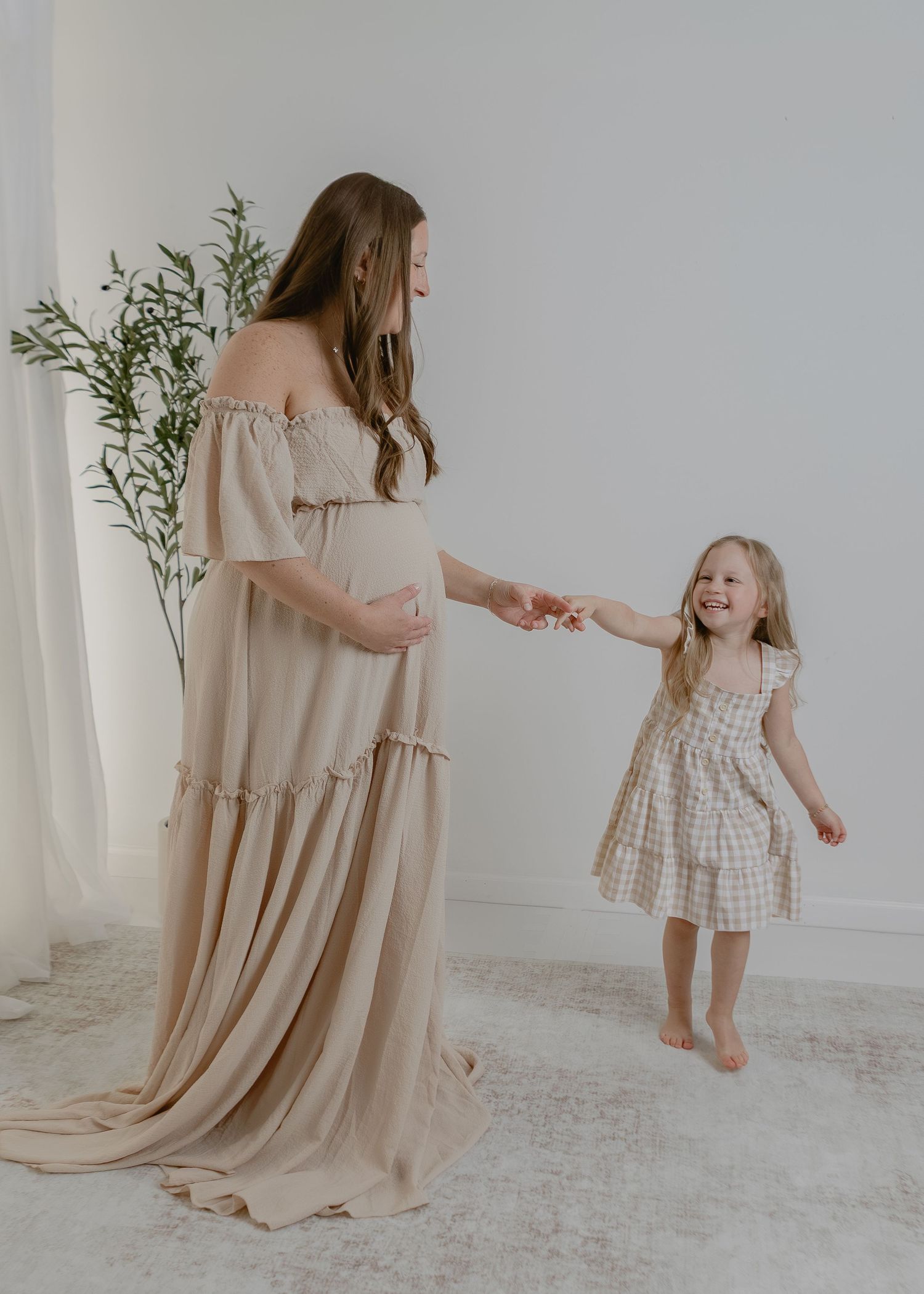 Pregnant mother in beige off-shoulder gown holding hands with her young daughter.
