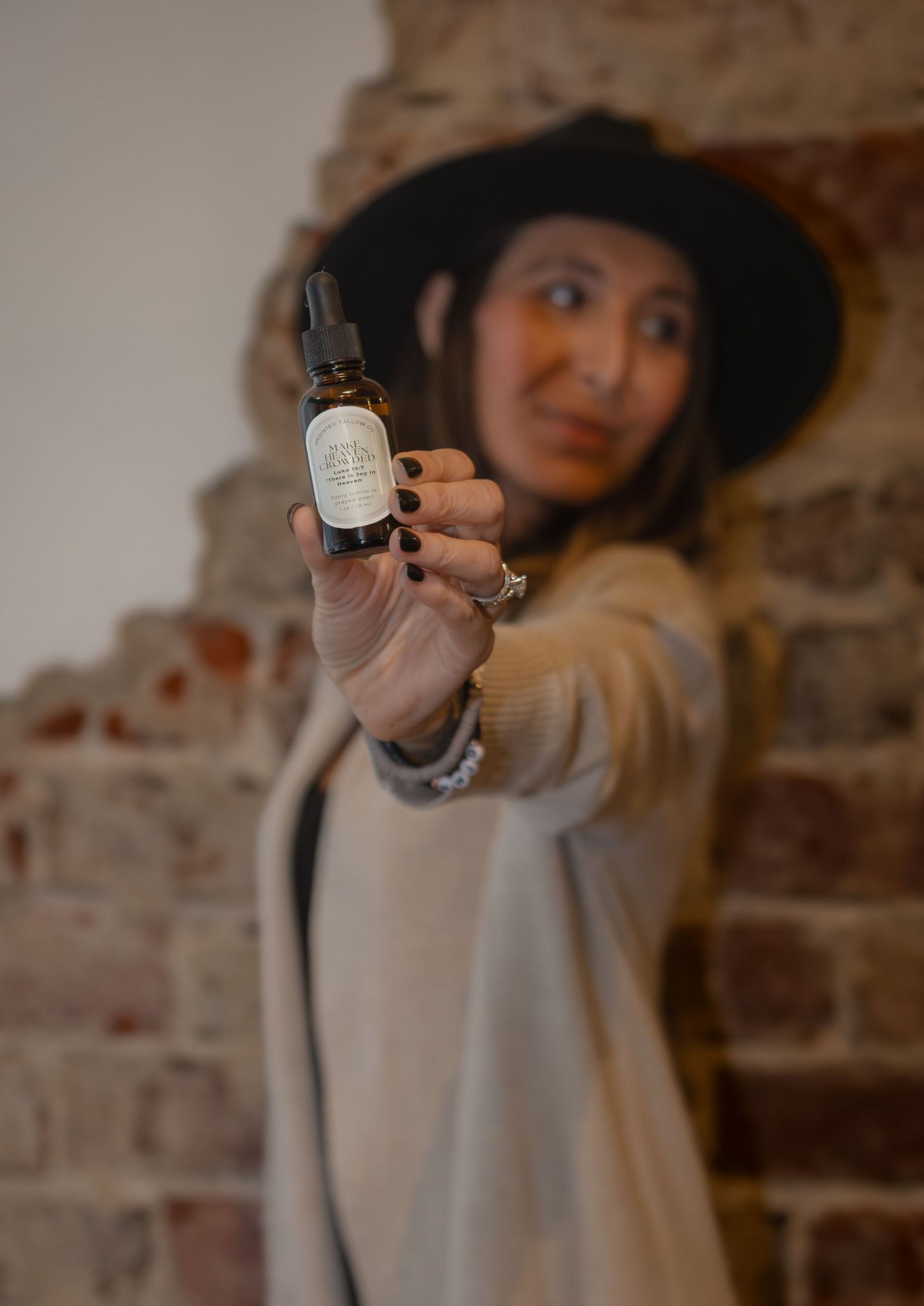 Woman in wide-brim hat holds small dropper bottle toward camera against an exposed brick wall background.