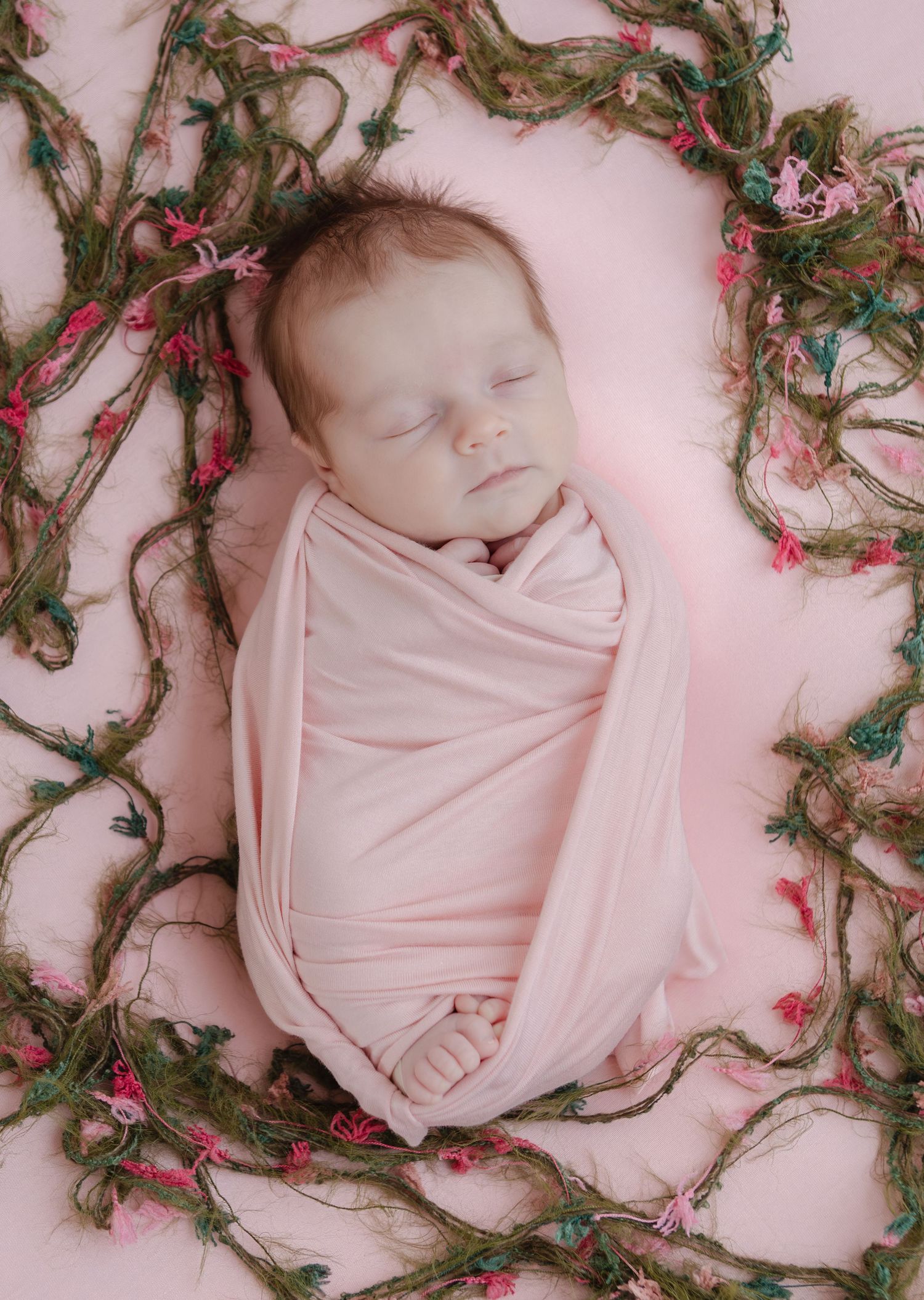 Sleeping newborn baby in pink swaddle wrap, nestled among green decorative vines on soft pink backdrop.