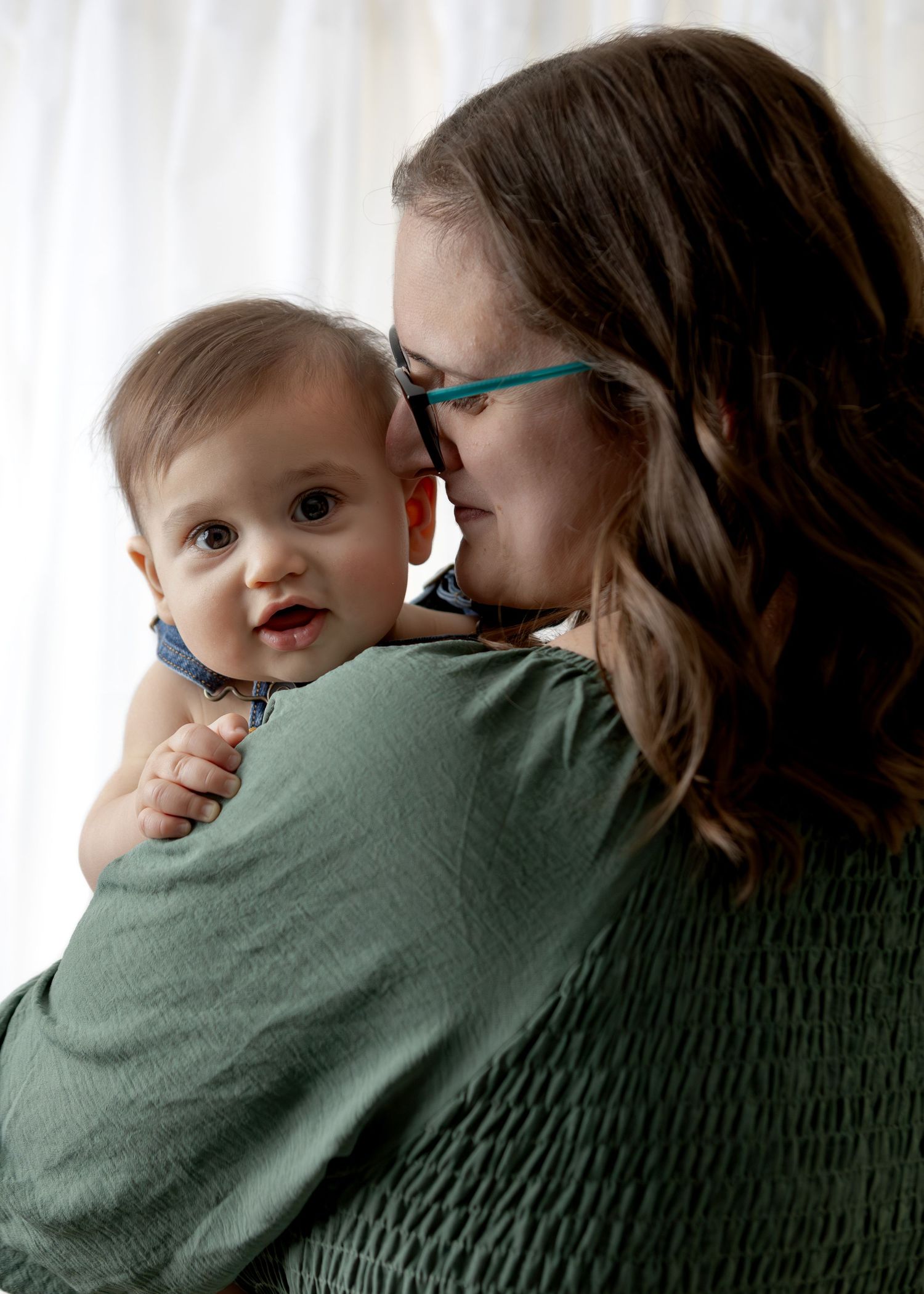 Mother with glasses in green top tenderly nuzzling her smiling baby boy indoors.