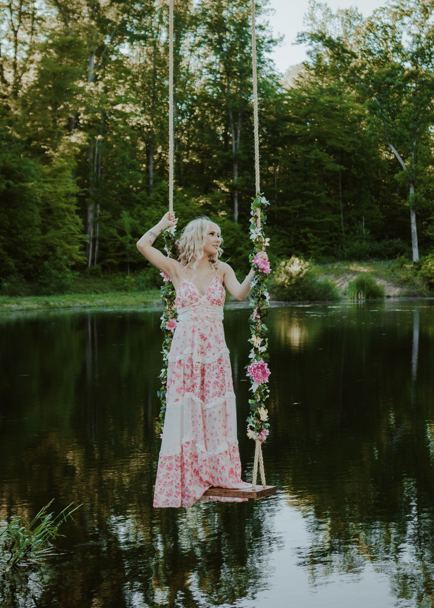 Blonde woman in pink floral maxi dress standing on flower-decorated swing over calm lake.