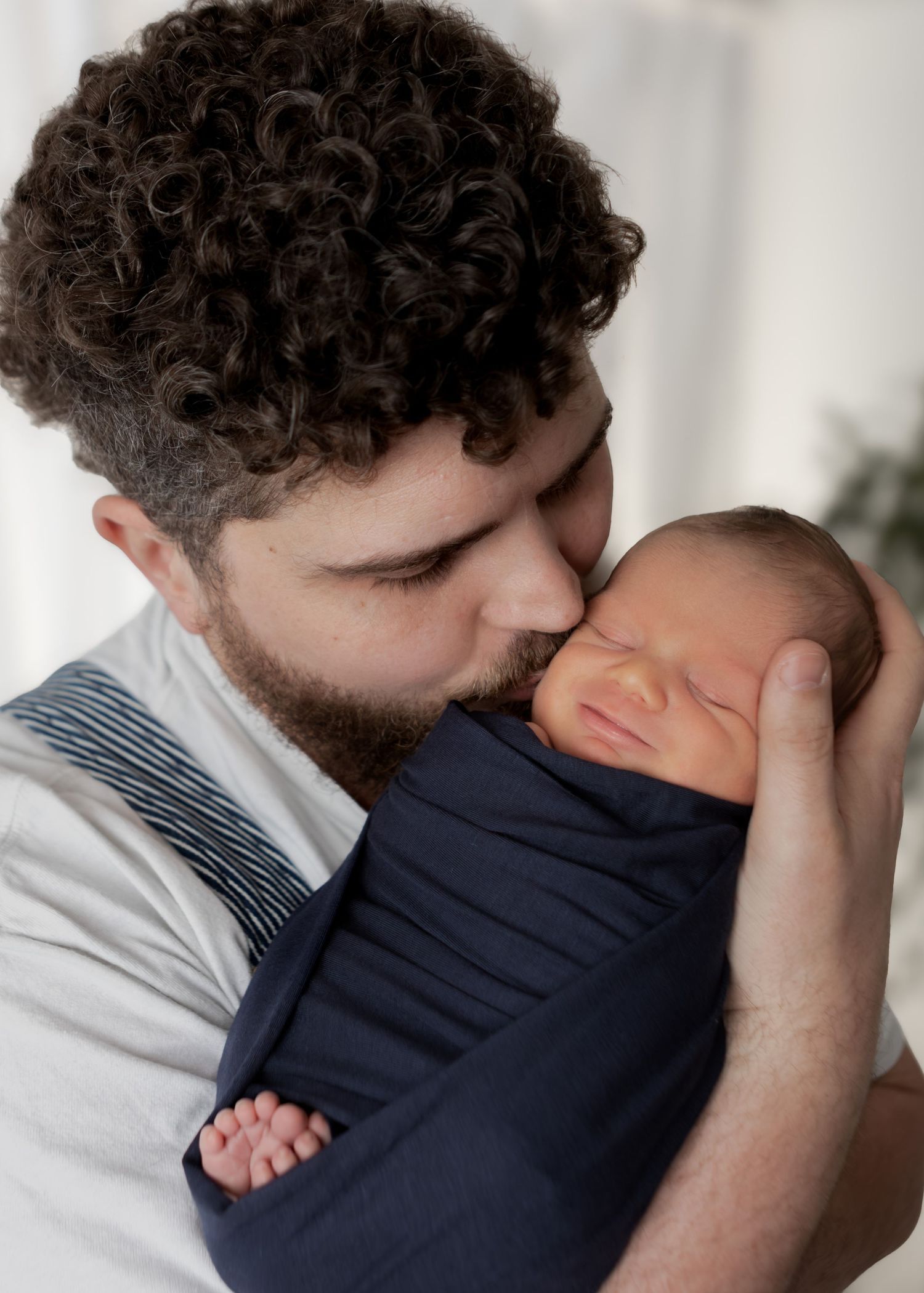 Curly-haired father tenderly holding and kissing his sleeping swaddled newborn baby.
