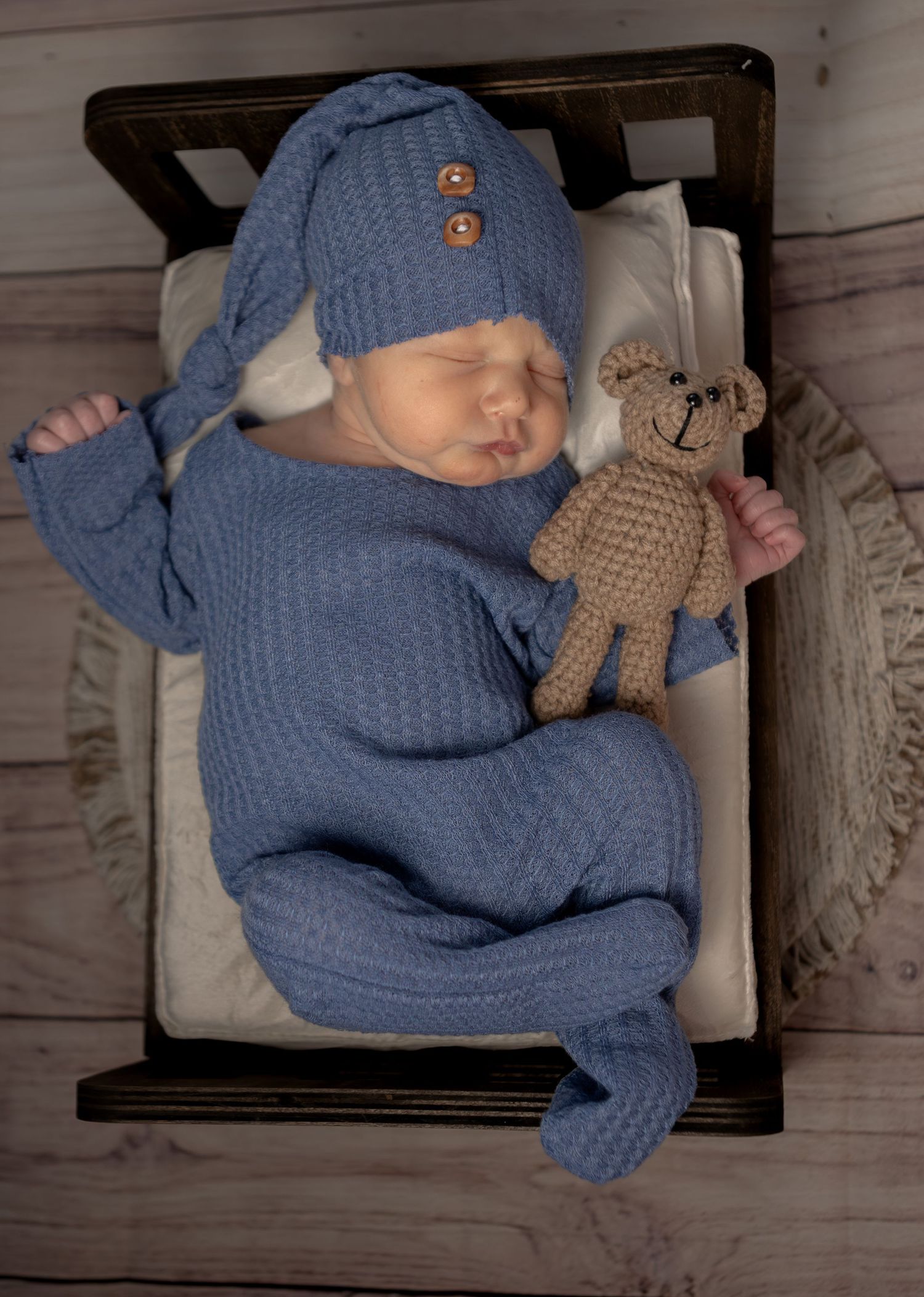 Sleeping newborn in blue knit outfit with matching hat holding a crocheted teddy bear.