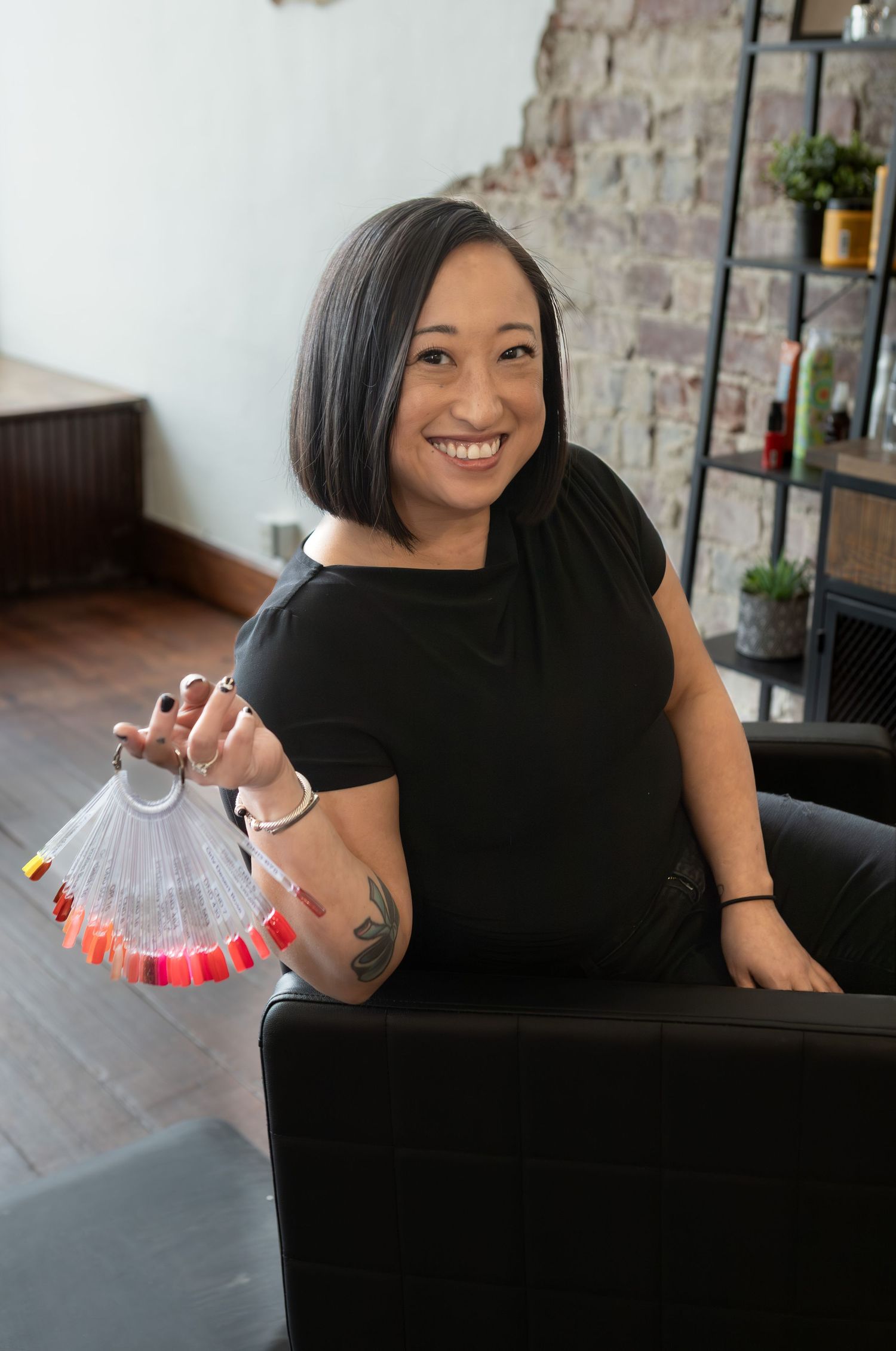 Smiling woman in black dress holds nail color swatch fan in a modern hair salon with brick wall.