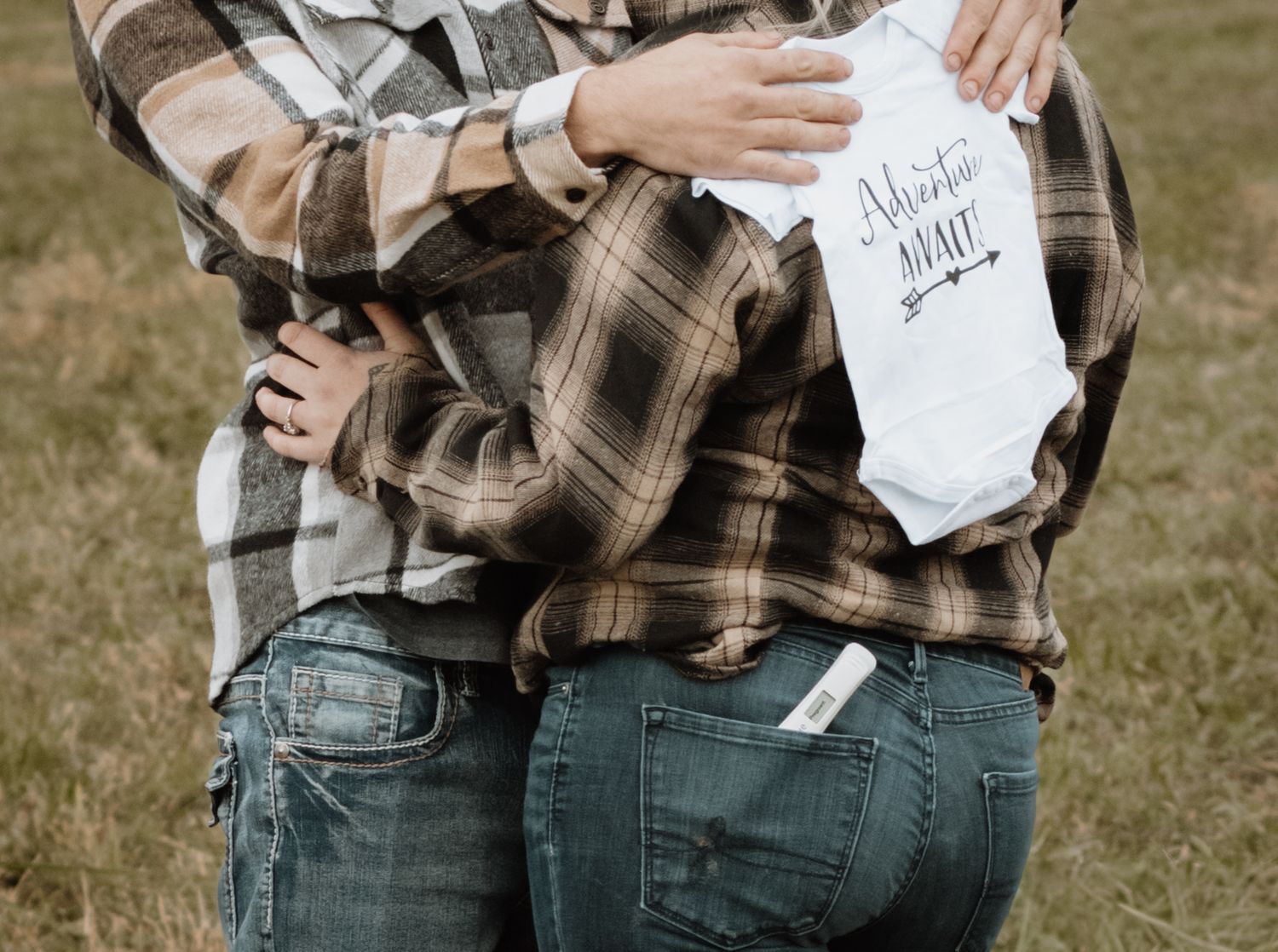 Couple in matching plaid shirts embracing outdoors, holding a white baby onesie announcing a pregnancy.