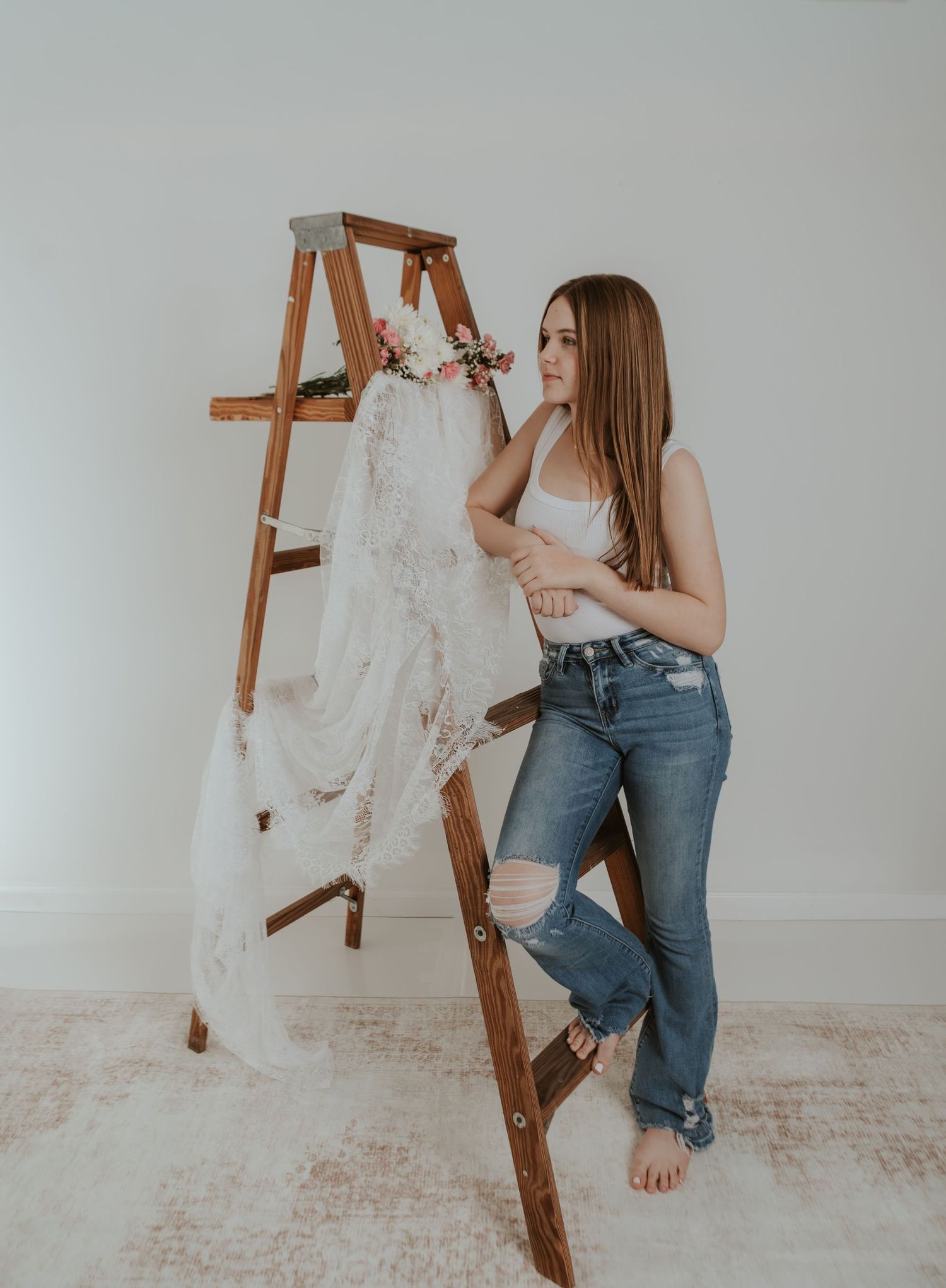 Young woman in white crop top and jeans leans against a wooden ladder displaying a lace wedding dress with flowers.