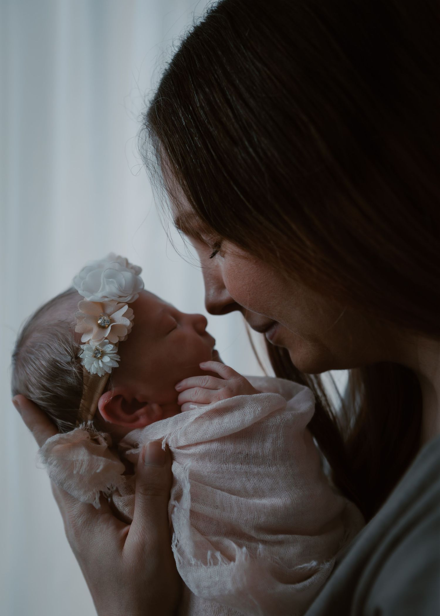 Mother tenderly holds her floral headband-adorned newborn baby girl face-to-face in soft light.