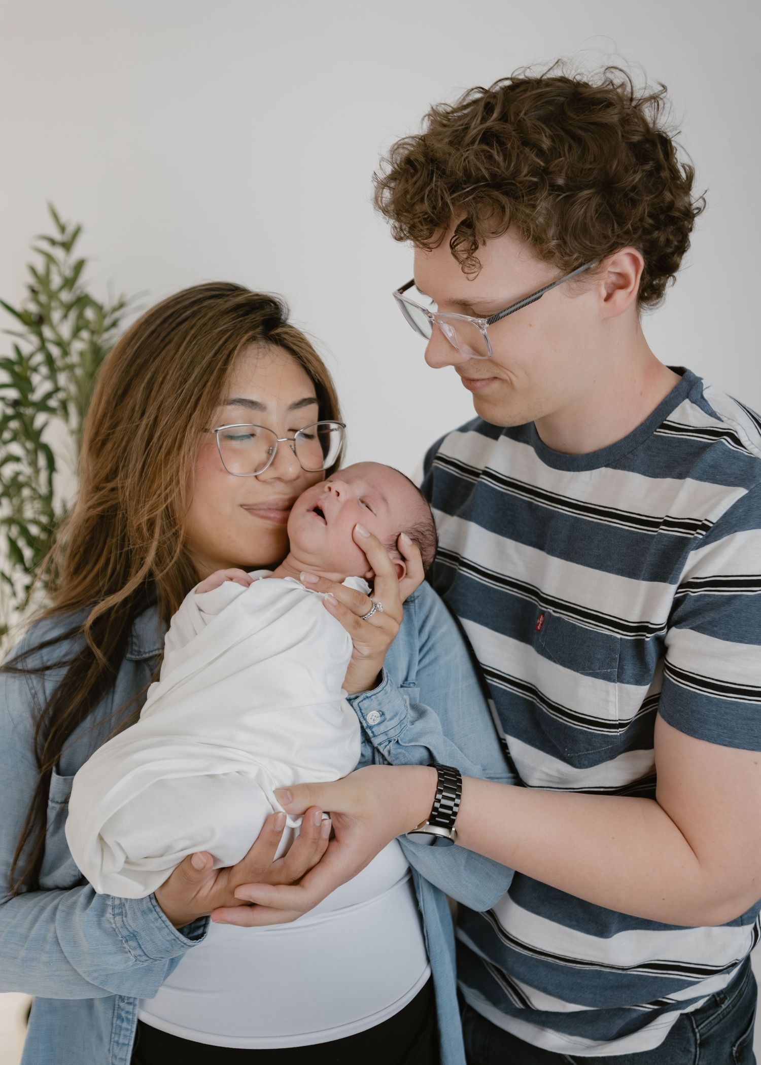 Young parents with glasses tenderly hold their swaddled newborn baby together indoors.