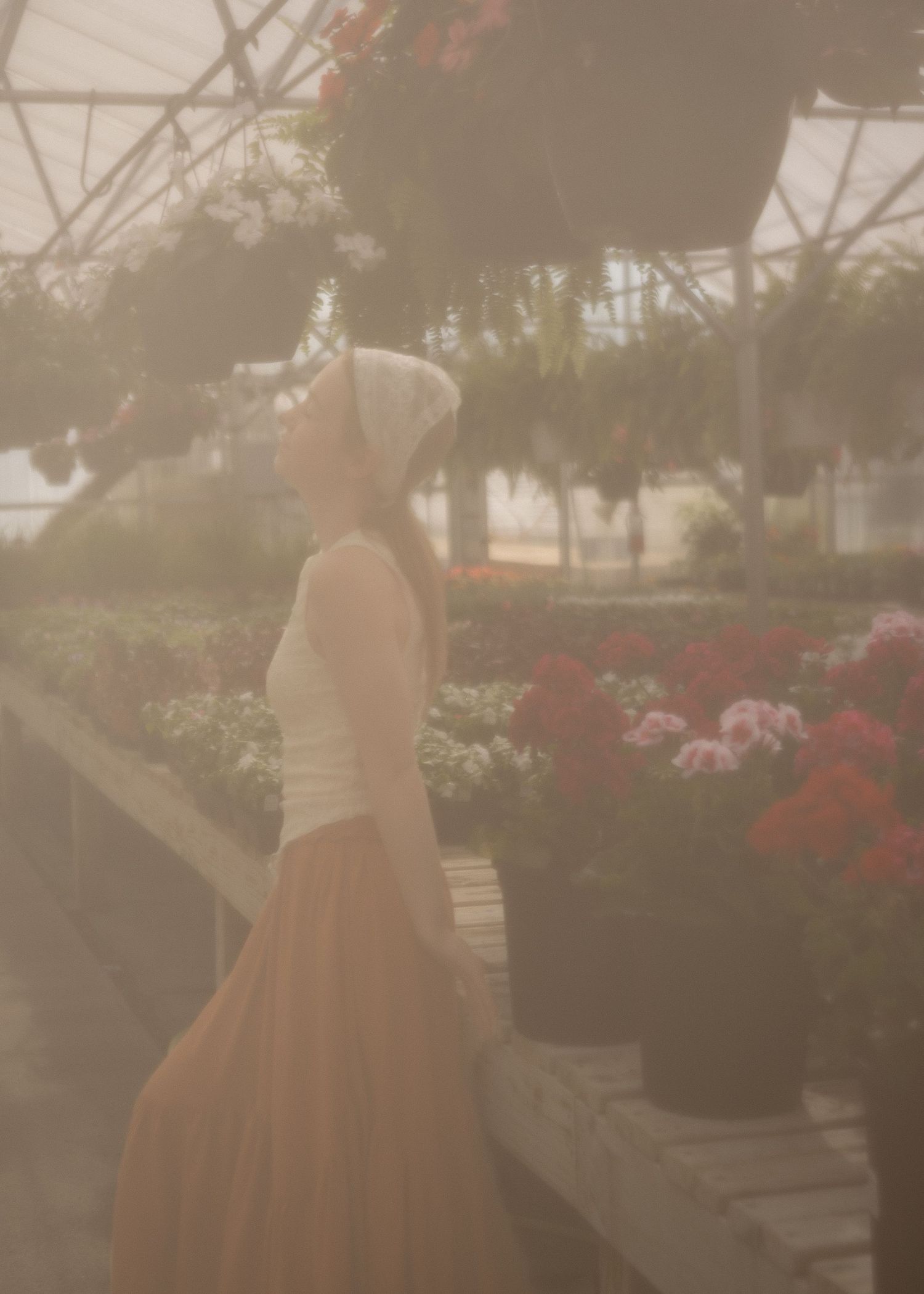 A woman in a white headscarf and rust skirt gazes upward among hanging flowers in a greenhouse.