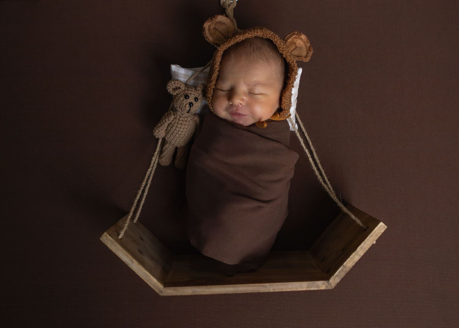 Smiling newborn wrapped in brown swaddle wearing bear bonnet in wooden hexagon swing prop.