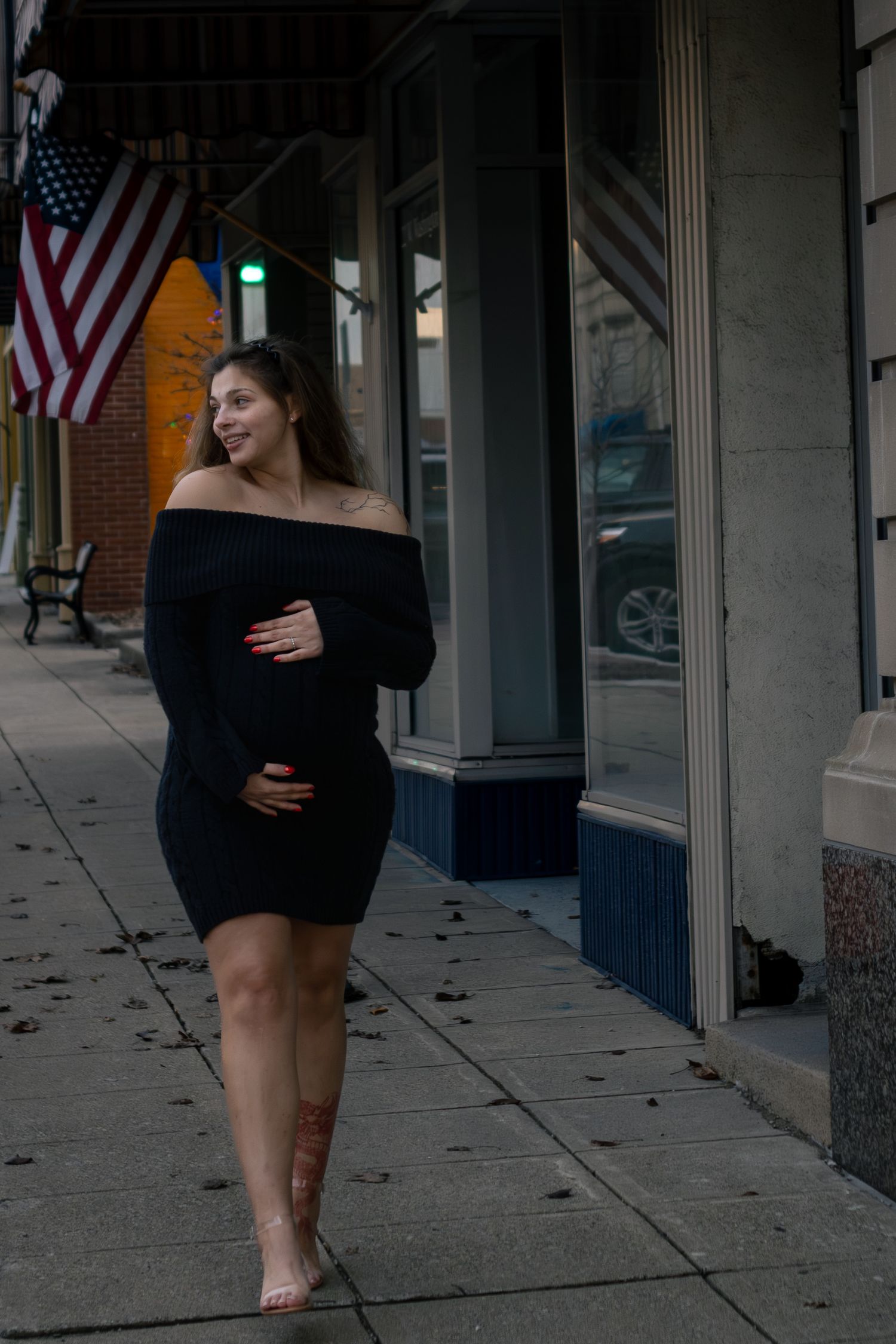 Smiling pregnant woman in black off-shoulder dress walking barefoot on a downtown sidewalk.