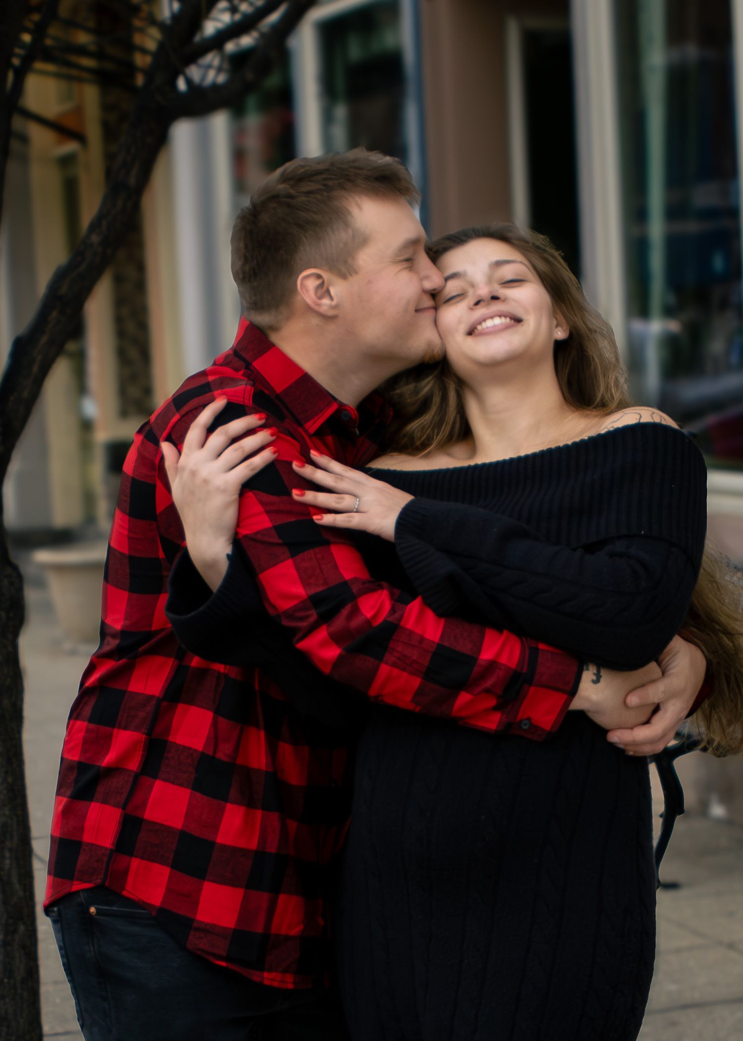 A couple shares a tender moment outdoors, man in red plaid shirt kissing smiling woman in black off-shoulder sweater.