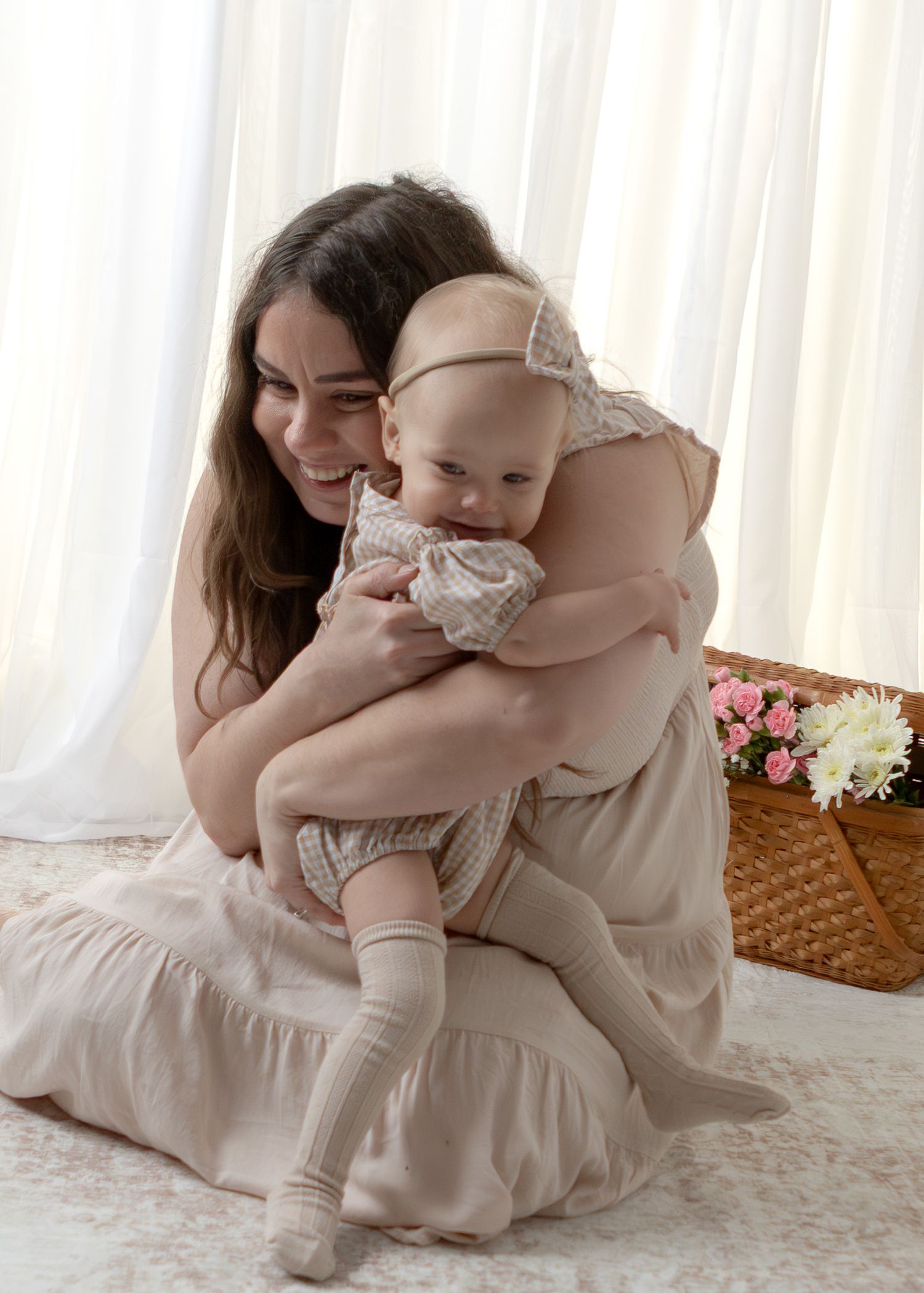 A brunette woman in a beige dress sits on the floor smiling while hugging her baby girl near a flower basket.