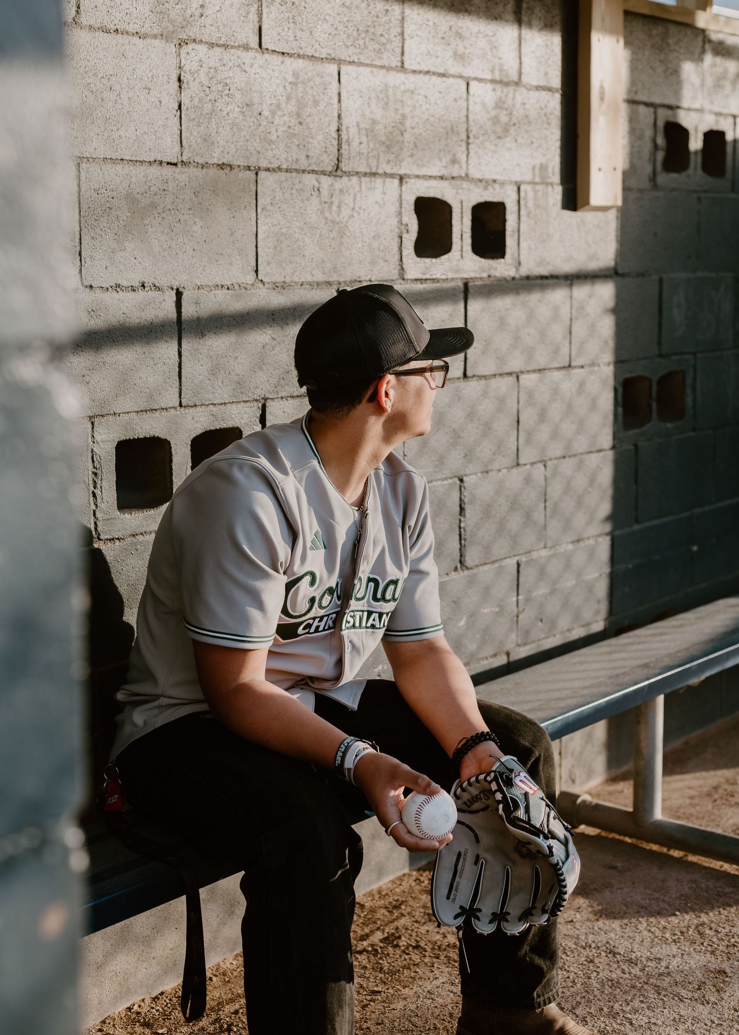 Male baseball player in Corona uniform sitting in dugout holding glove, wearing cap and glasses against brick wall.