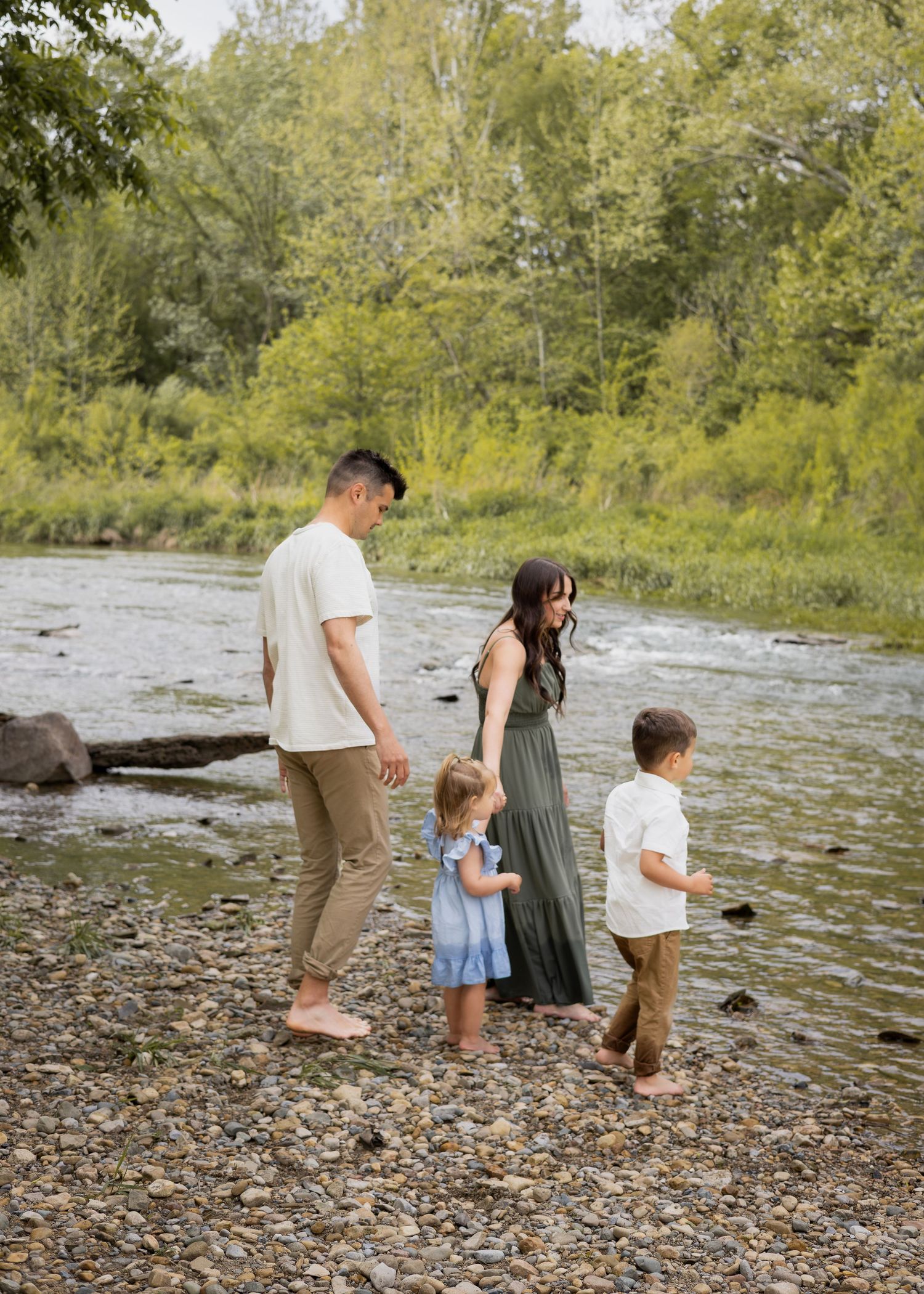 Family of four walking barefoot along a rocky riverbank surrounded by lush green trees.
