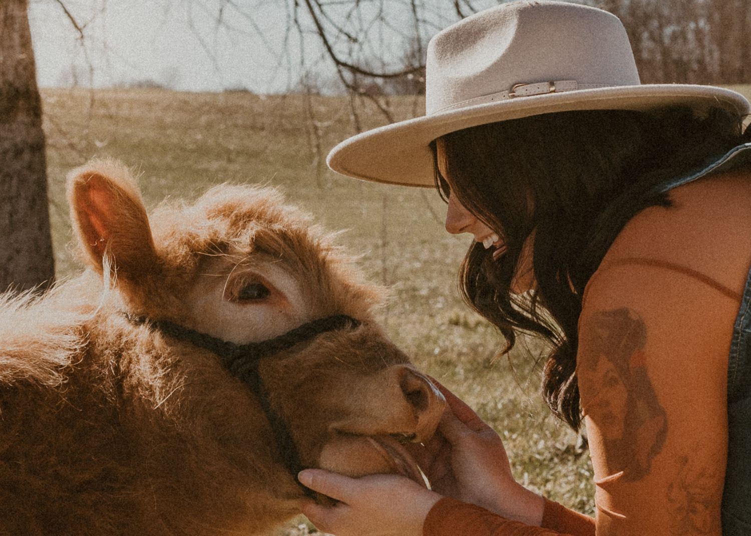 Woman in wide-brim hat tenderly petting a fluffy brown miniature Highland calf outdoors.