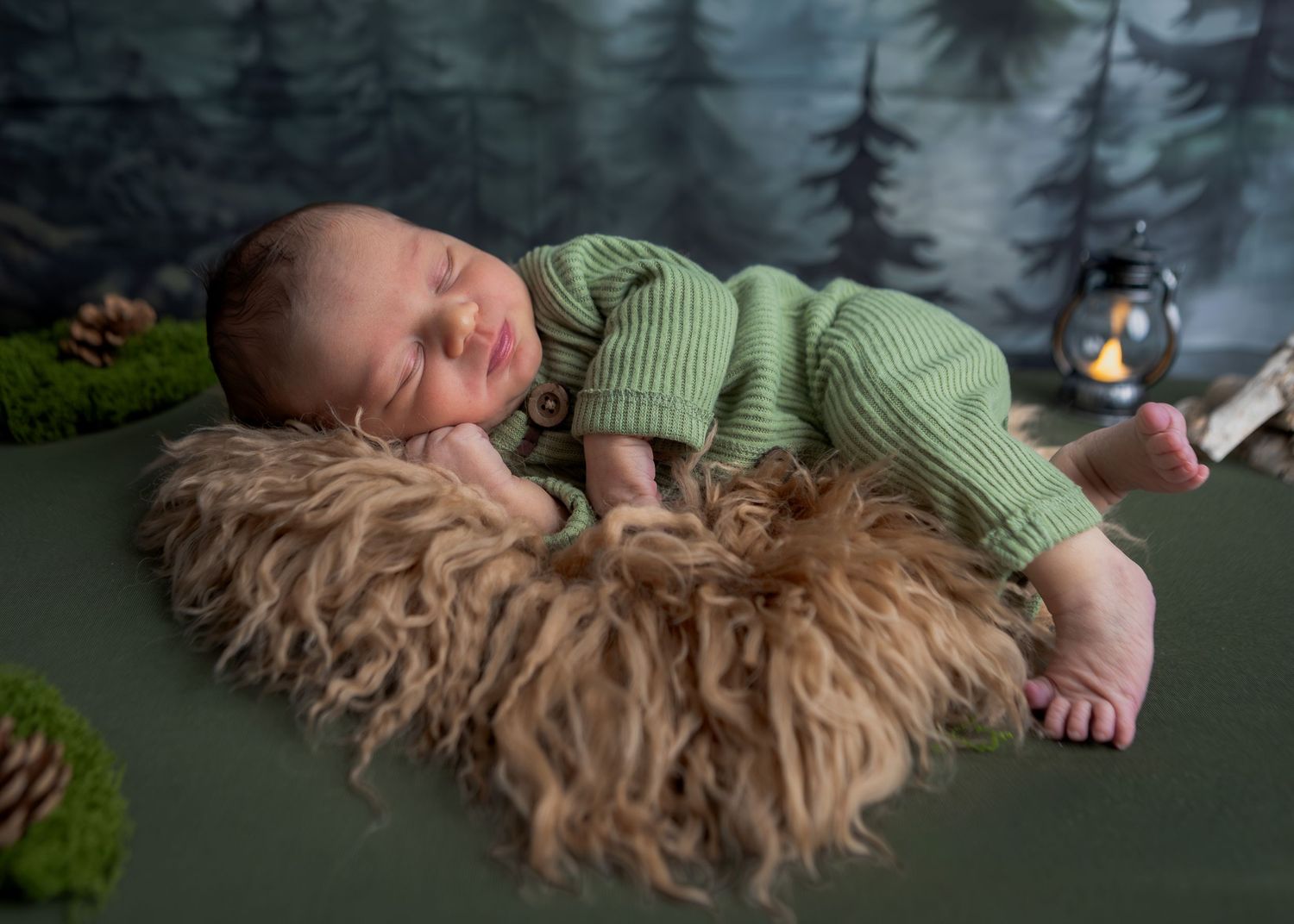 Sleeping newborn baby in green romper posed on fluffy fur in woodland-themed photo setup.