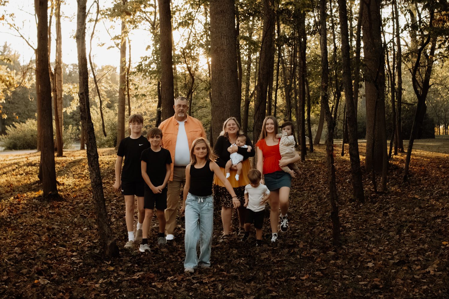 Large family of eight posing among autumn trees with golden sunlight filtering through the forest.