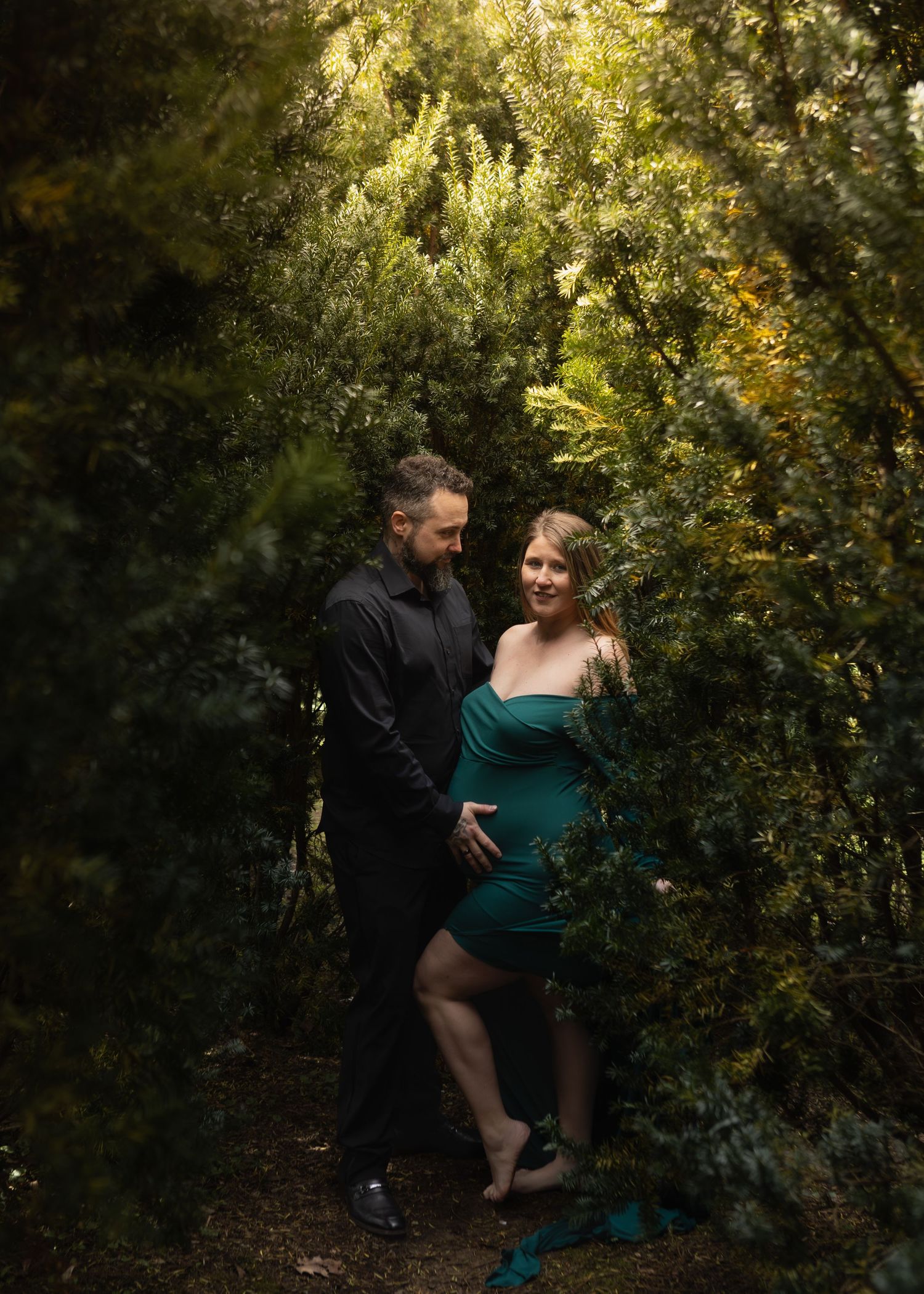 Expecting couple in teal and black standing together surrounded by lush green foliage outdoors.