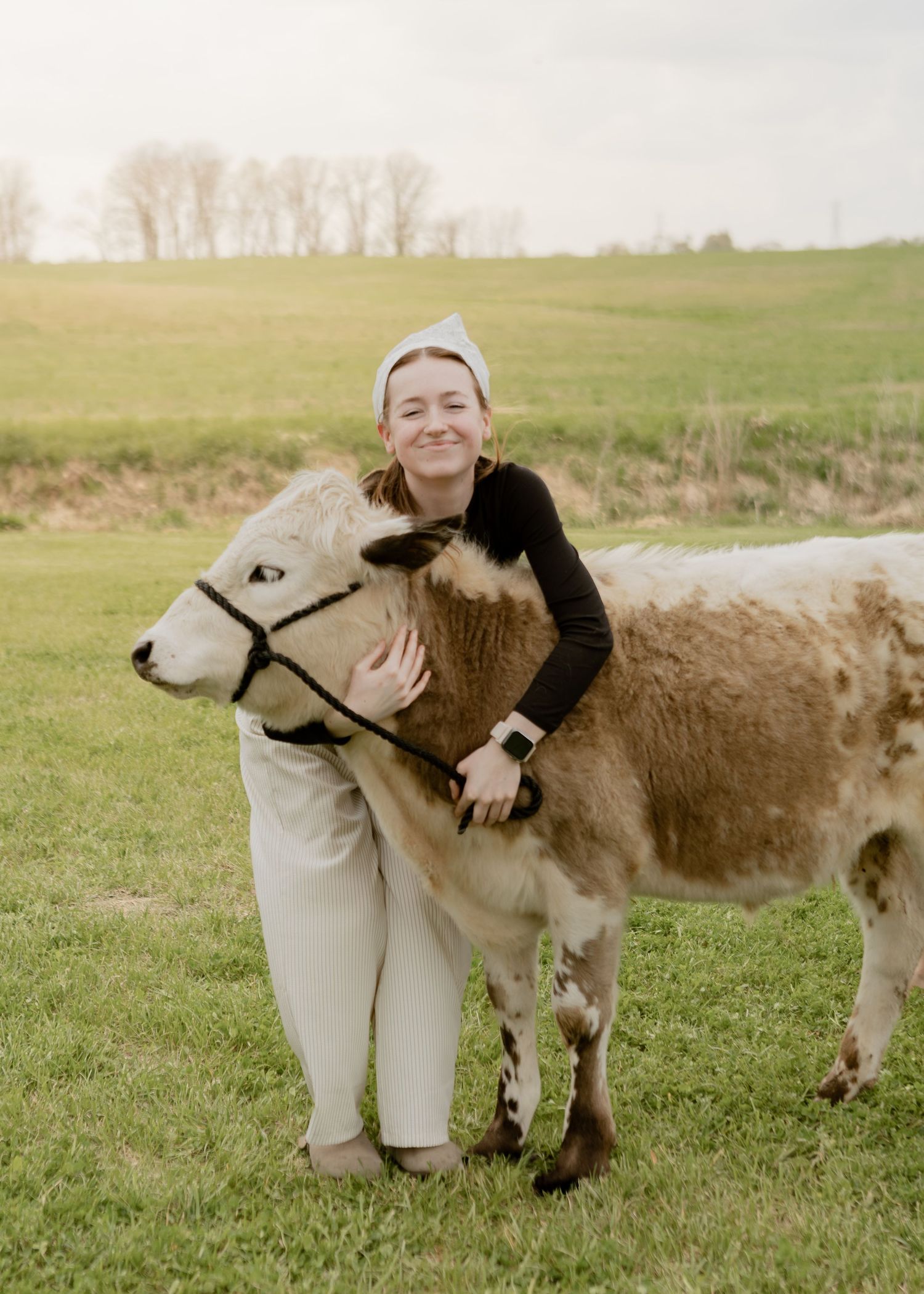 Smiling woman hugging a brown and white miniature calf while standing in a green open field.
