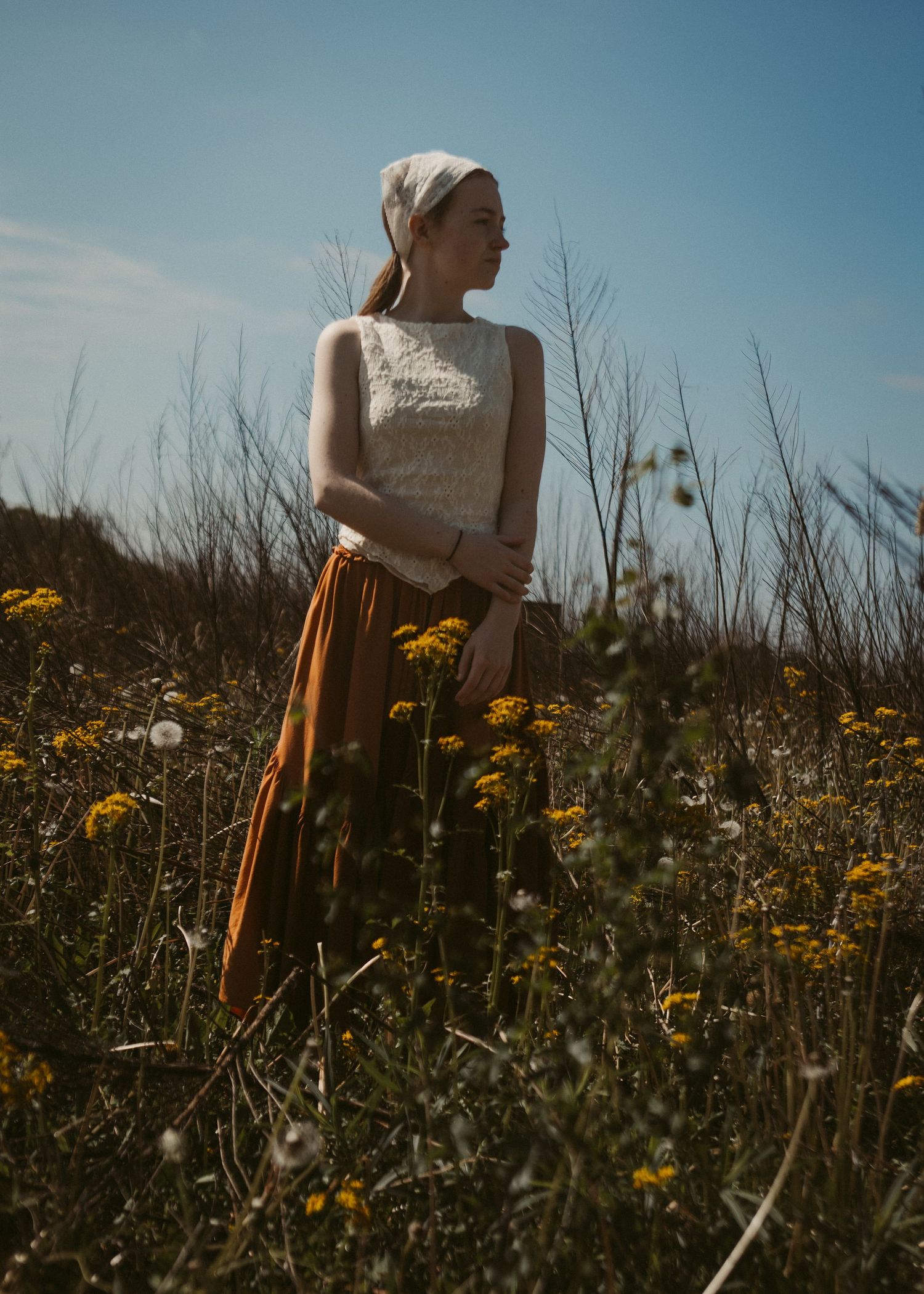 A young woman in a white top and rust skirt stands in a wildflower field under blue sky.
