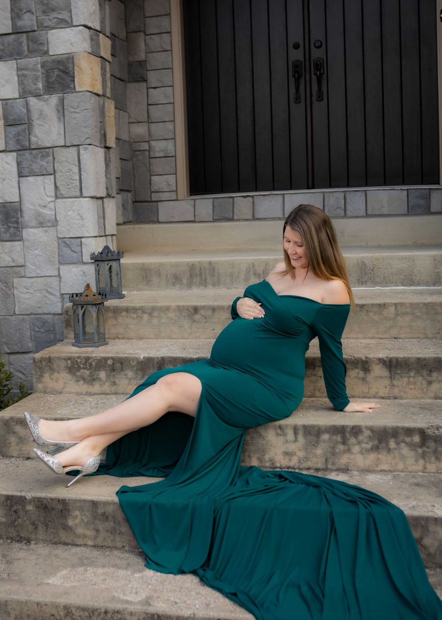 Pregnant woman in elegant emerald gown sitting on stone steps outside castle-style home.