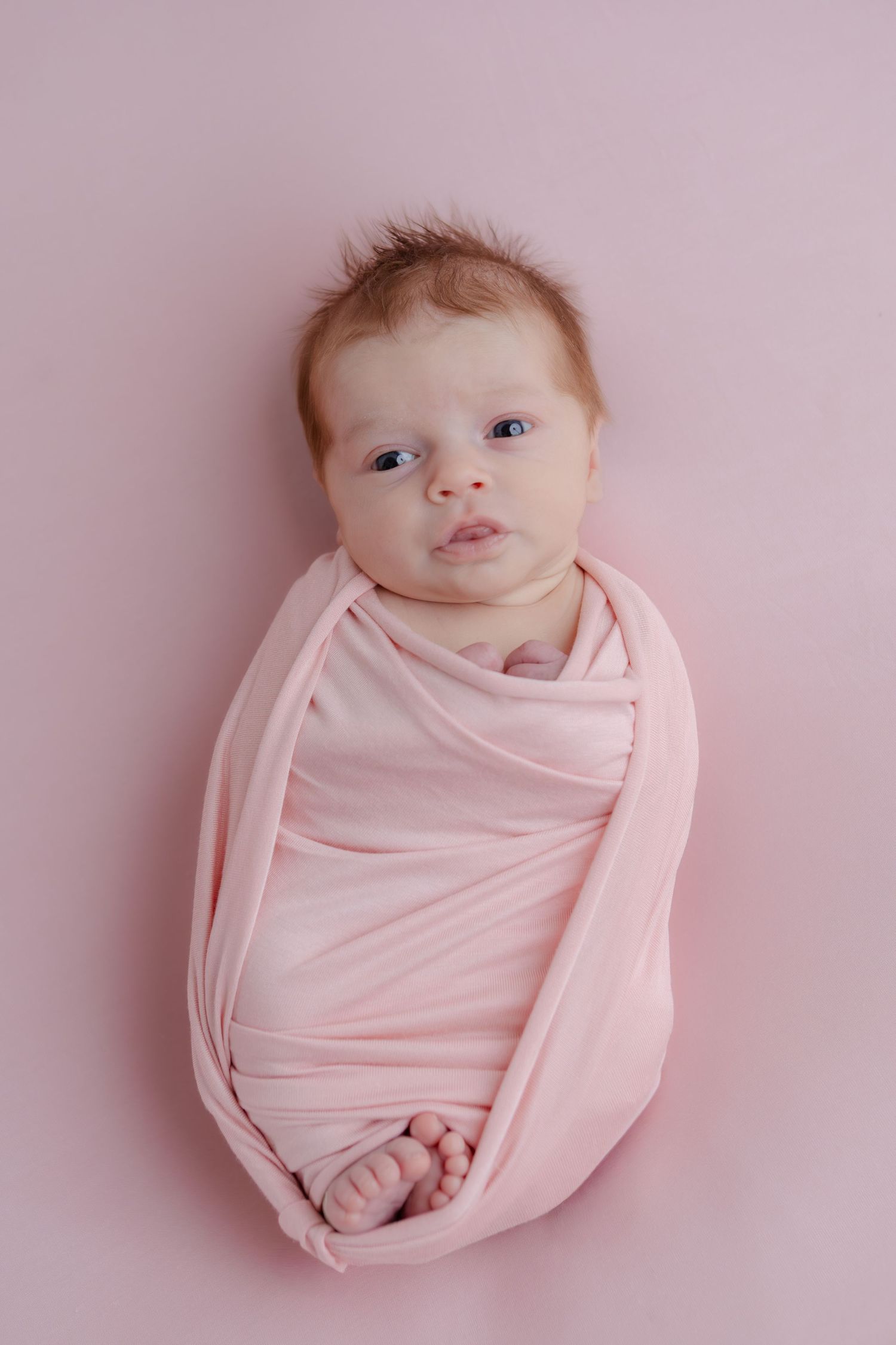 Newborn baby wrapped snugly in pink swaddle blanket with tiny toes peeking out on pink backdrop.