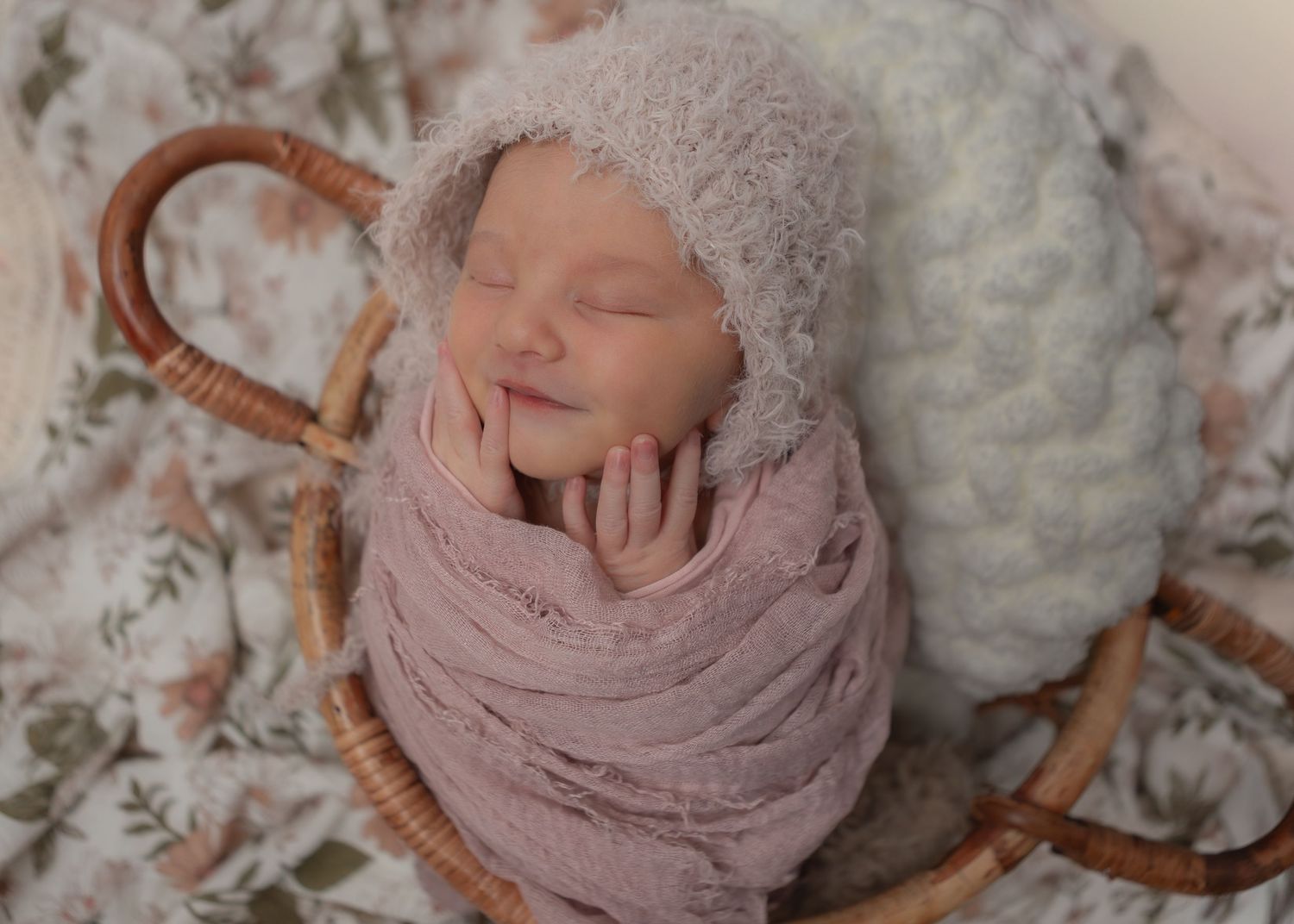 Sleeping newborn baby in mauve wrap and fuzzy bonnet posed in wicker basket.