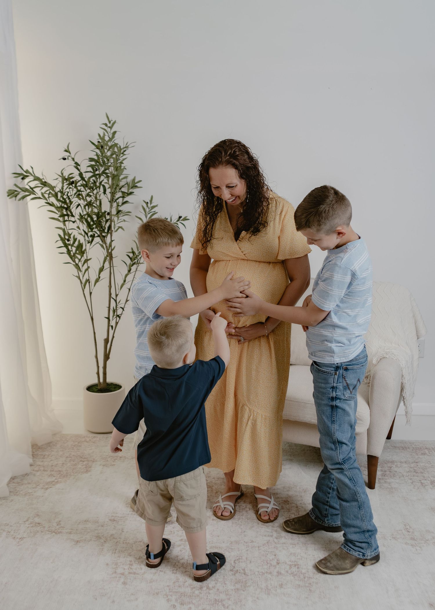 Pregnant woman in yellow dress smiling as three young boys touch her baby bump indoors.