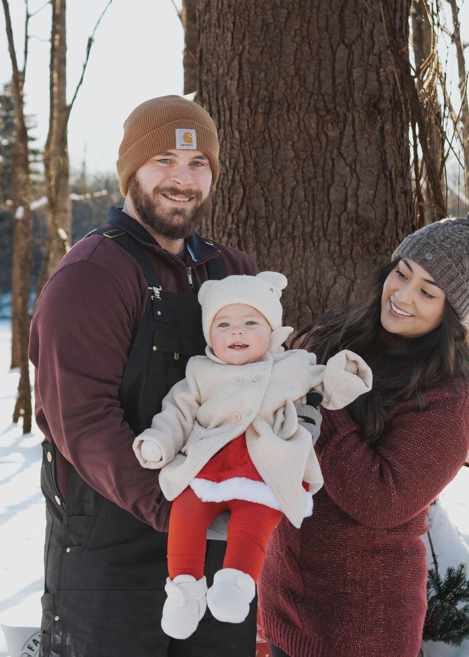 A young couple in winter attire holds their smiling baby dressed in a festive red outfit outdoors in the snow.