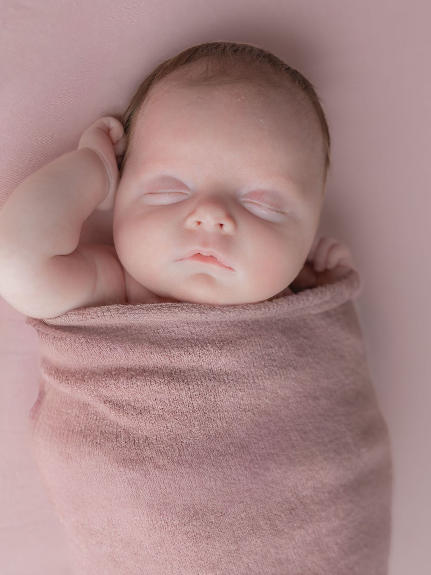 Peaceful sleeping newborn baby swaddled in a soft pink wrap with hand raised beside face.