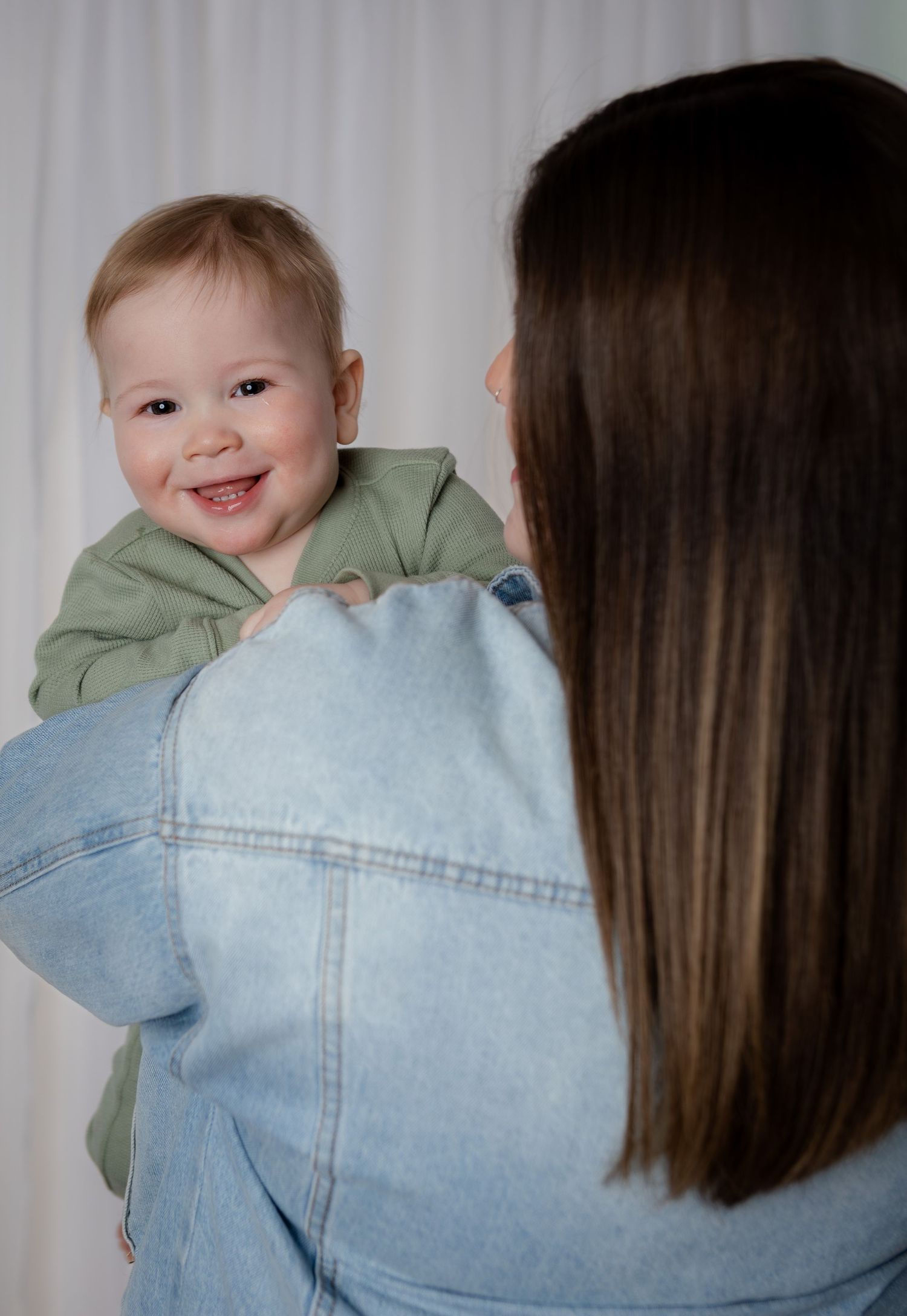 Smiling baby in green hoodie looks over mothers shoulder as she holds him in a bright studio.