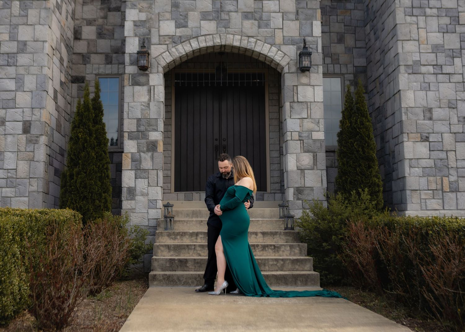 Couple embracing before a stone castle-style building, woman in emerald green gown.