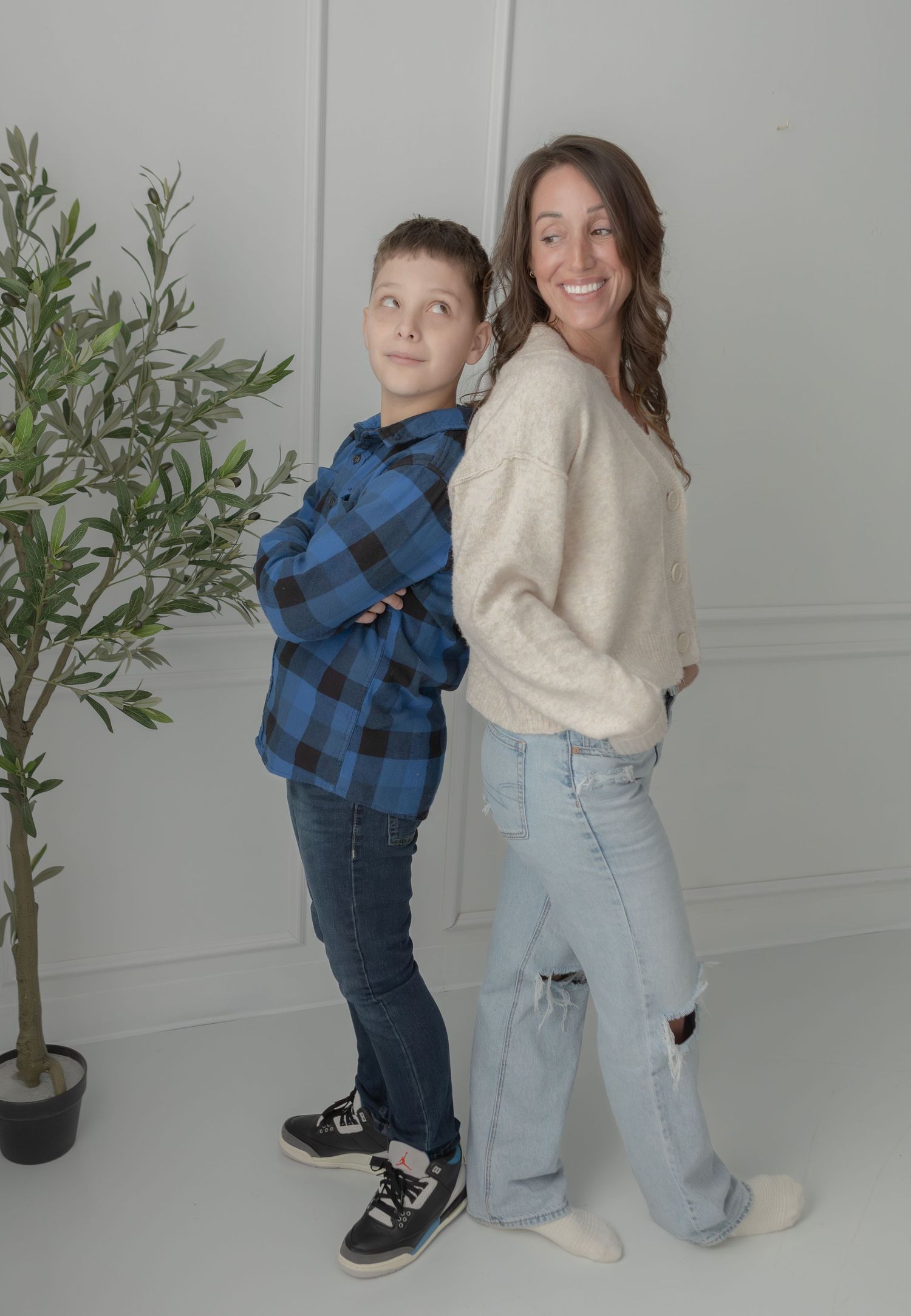 Smiling mother and preteen son pose back-to-back in a bright white studio near an olive tree.