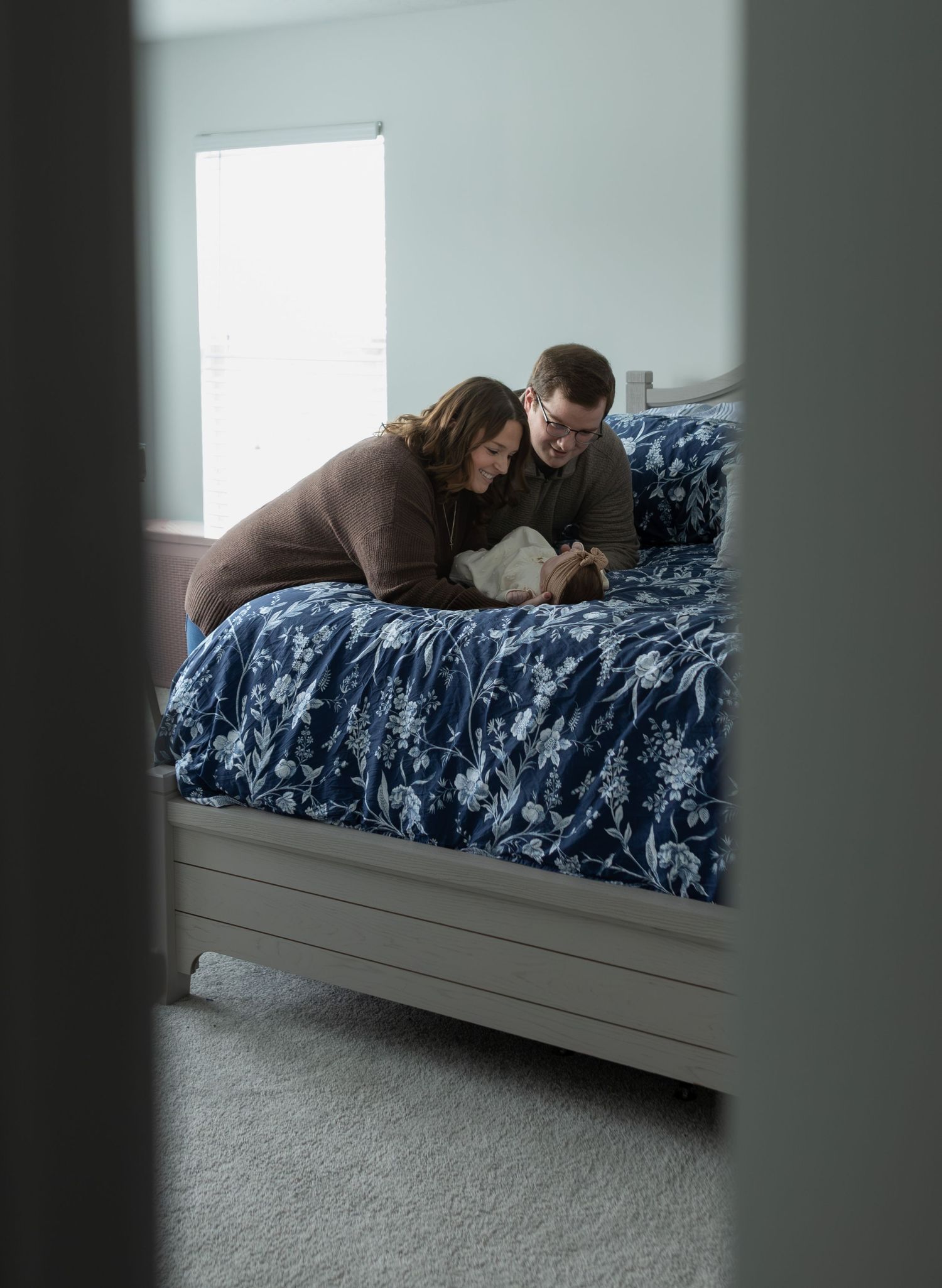 A couple leans over their newborn baby on a blue floral bedspread in a warmly lit bedroom.
