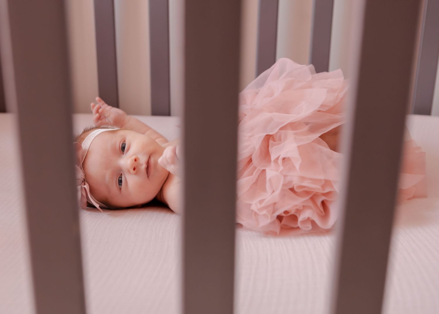 Adorable baby girl in a pink tutu lying in a wooden crib looking at the camera.