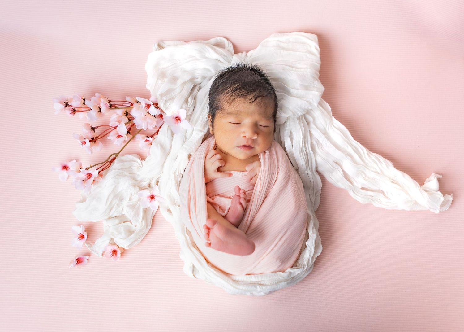 Peacefully sleeping newborn wrapped in pink swaddle beside cherry blossoms on pink backdrop.