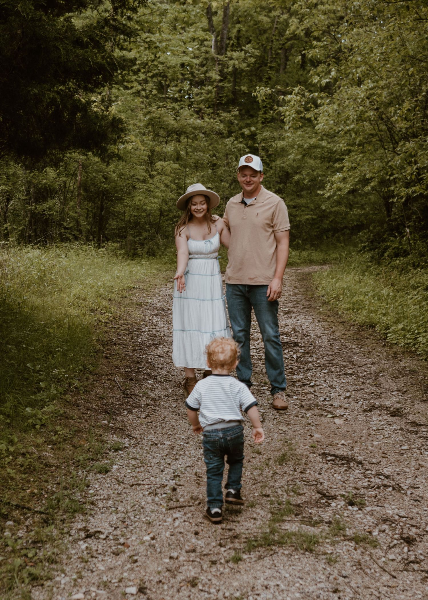 Young family walking on wooded trail, toddler running toward smiling parents in natural outdoor setting.