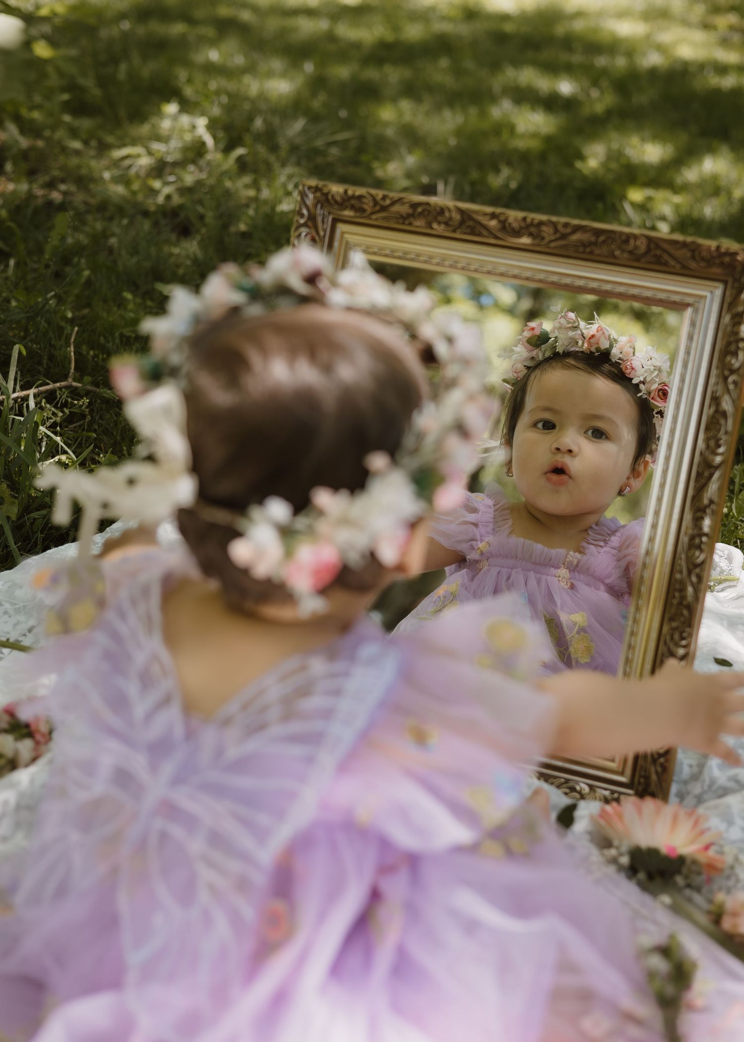 Adorable baby girl in purple fairy dress and floral crown looking into ornate mirror.