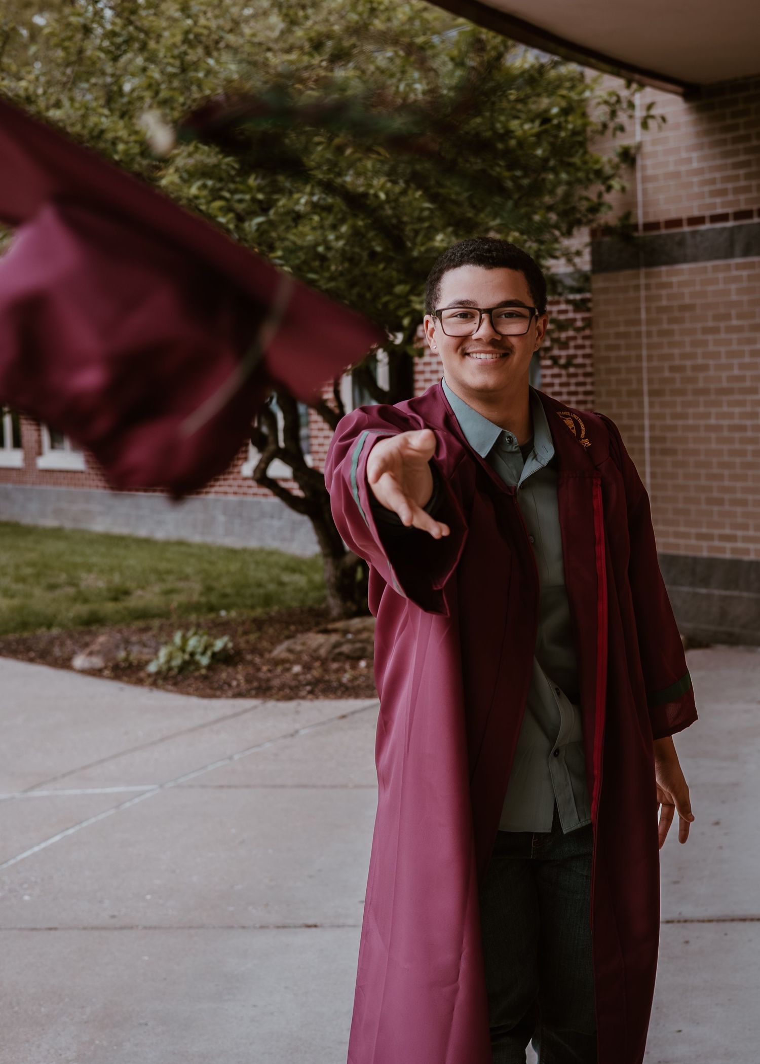 A smiling graduate in a maroon gown reaches toward the camera outside a brick school building.