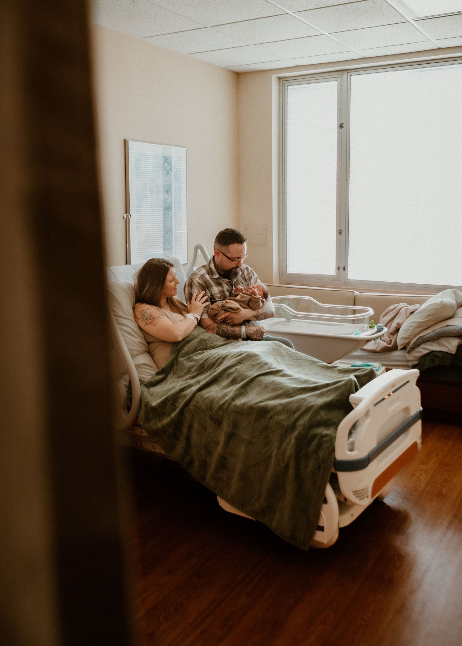 New parents sit together on hospital bed looking at their newborn baby by a bright window.