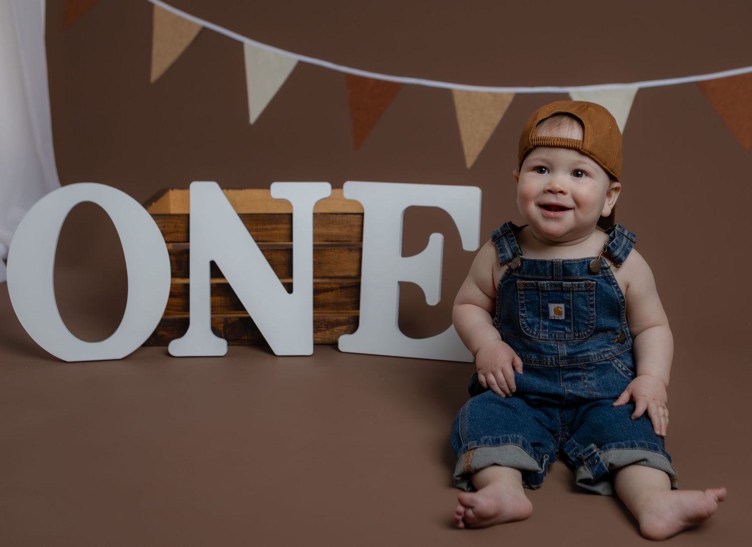 Happy one-year-old boy in denim overalls and brown cap sitting in front of ONE letters for birthday photos.