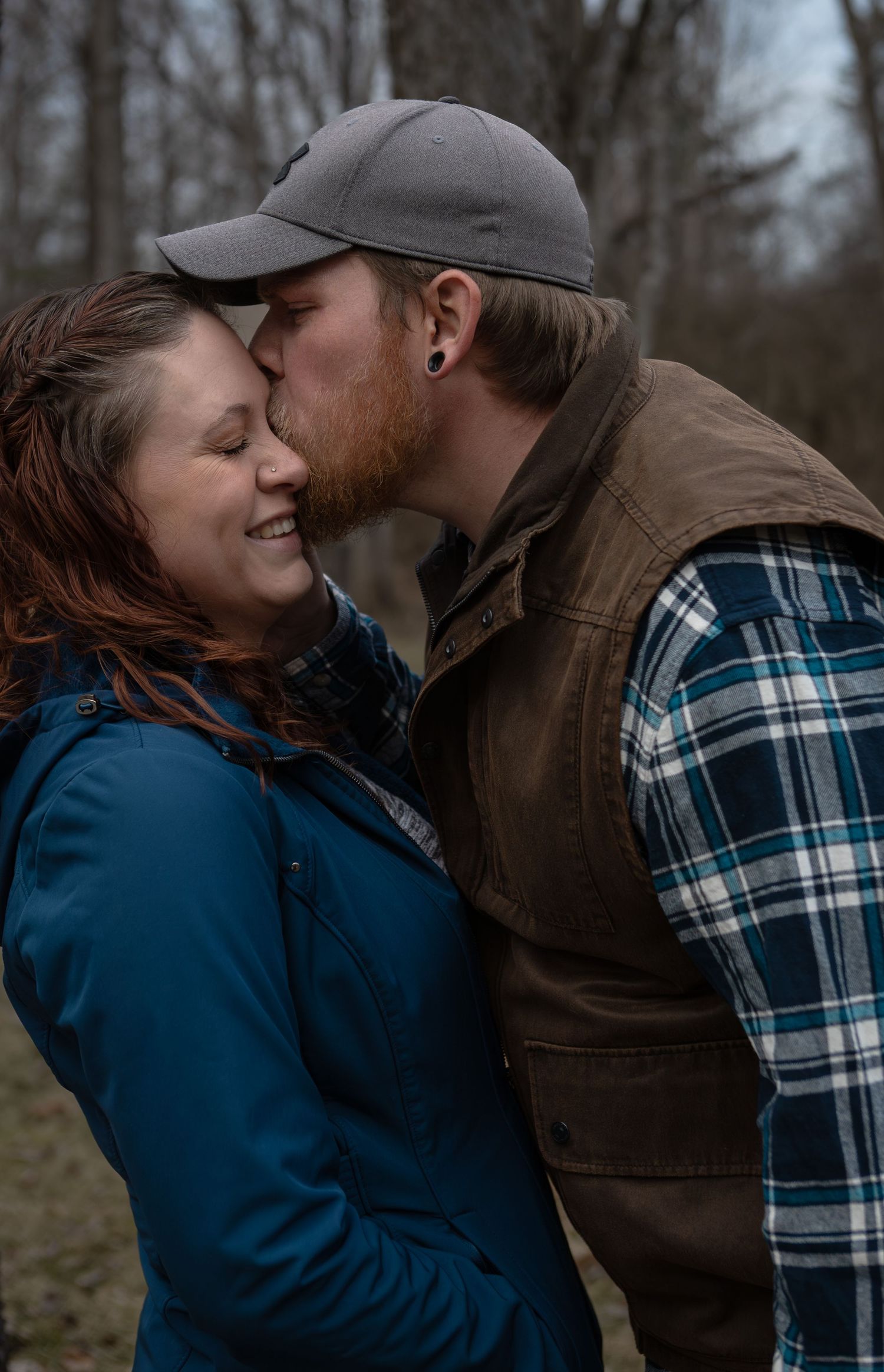 Couple sharing a tender kiss outdoors, wearing casual flannel and jacket in a wooded setting.