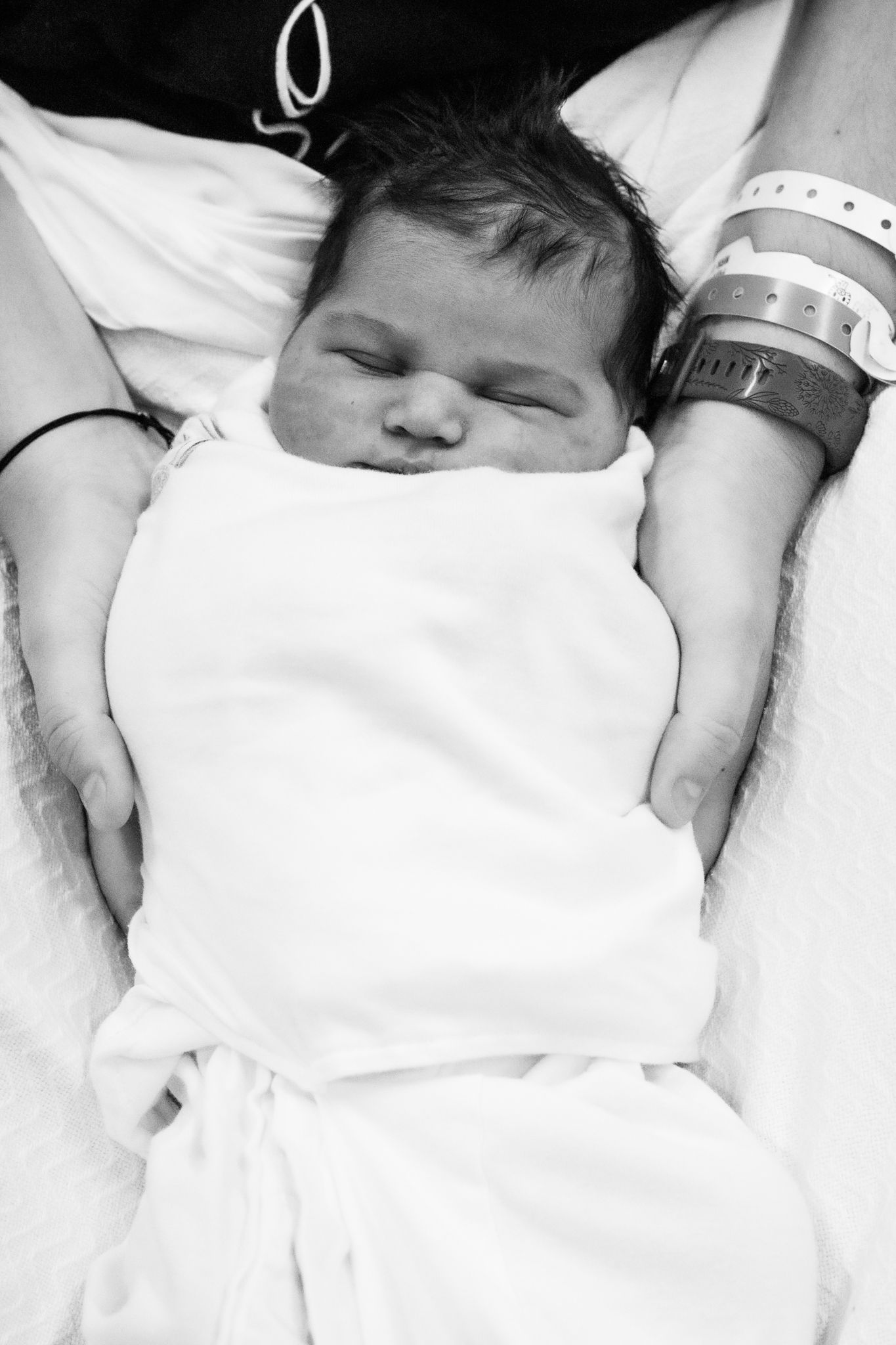 Black and white close-up of a sleeping swaddled newborn resting in a parent's arms wearing a hospital bracelet.