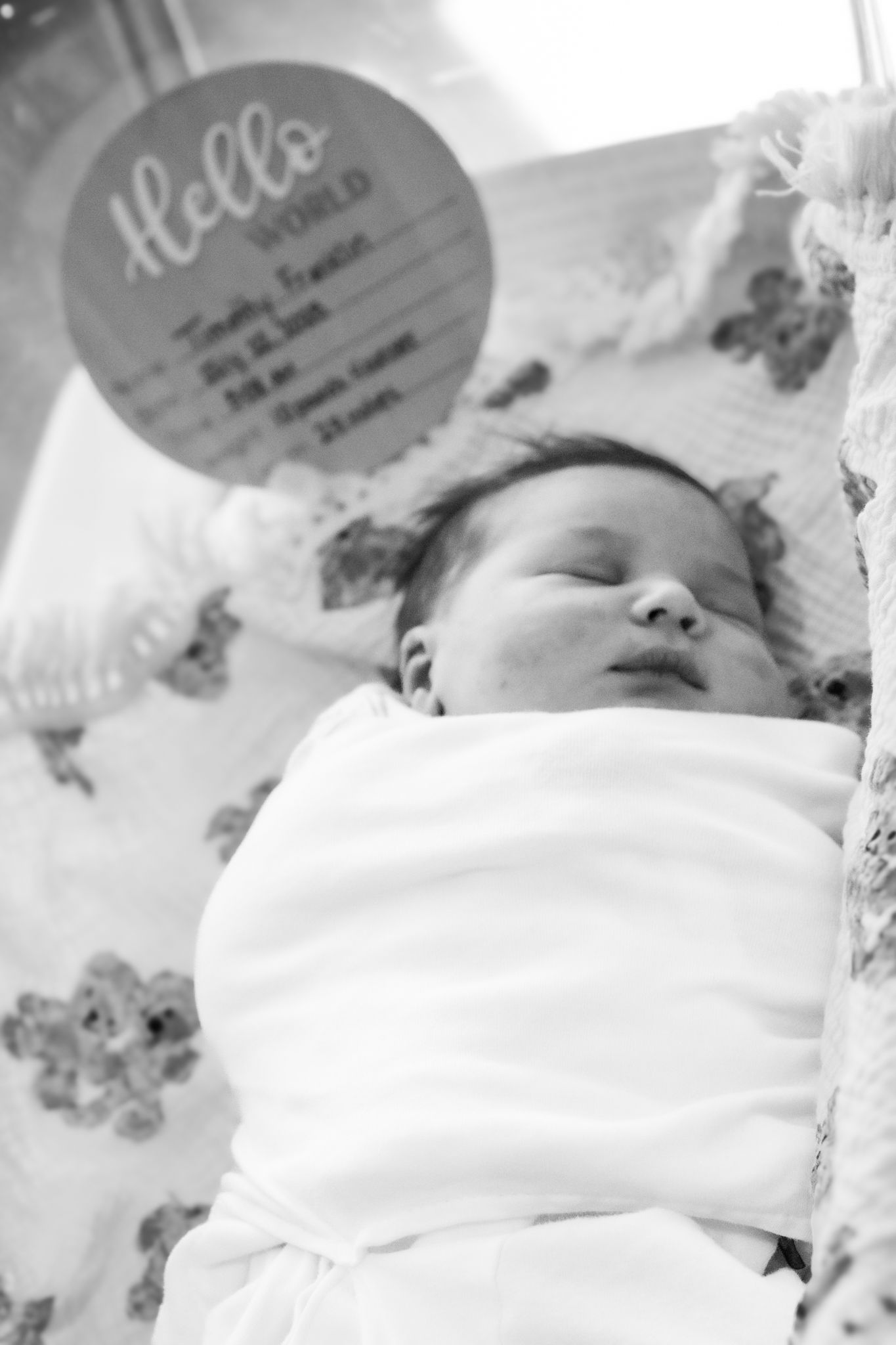 Sleeping newborn baby swaddled in white blanket with Hello World birth announcement sign in hospital.
