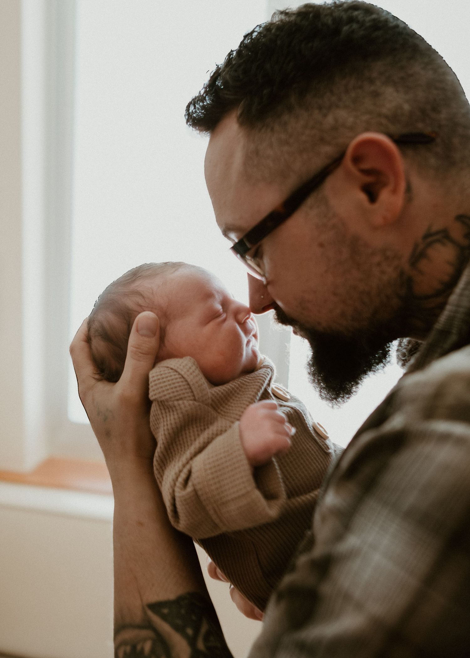 Tattooed father with glasses gently holds and kisses his sleeping newborn baby near a bright window.