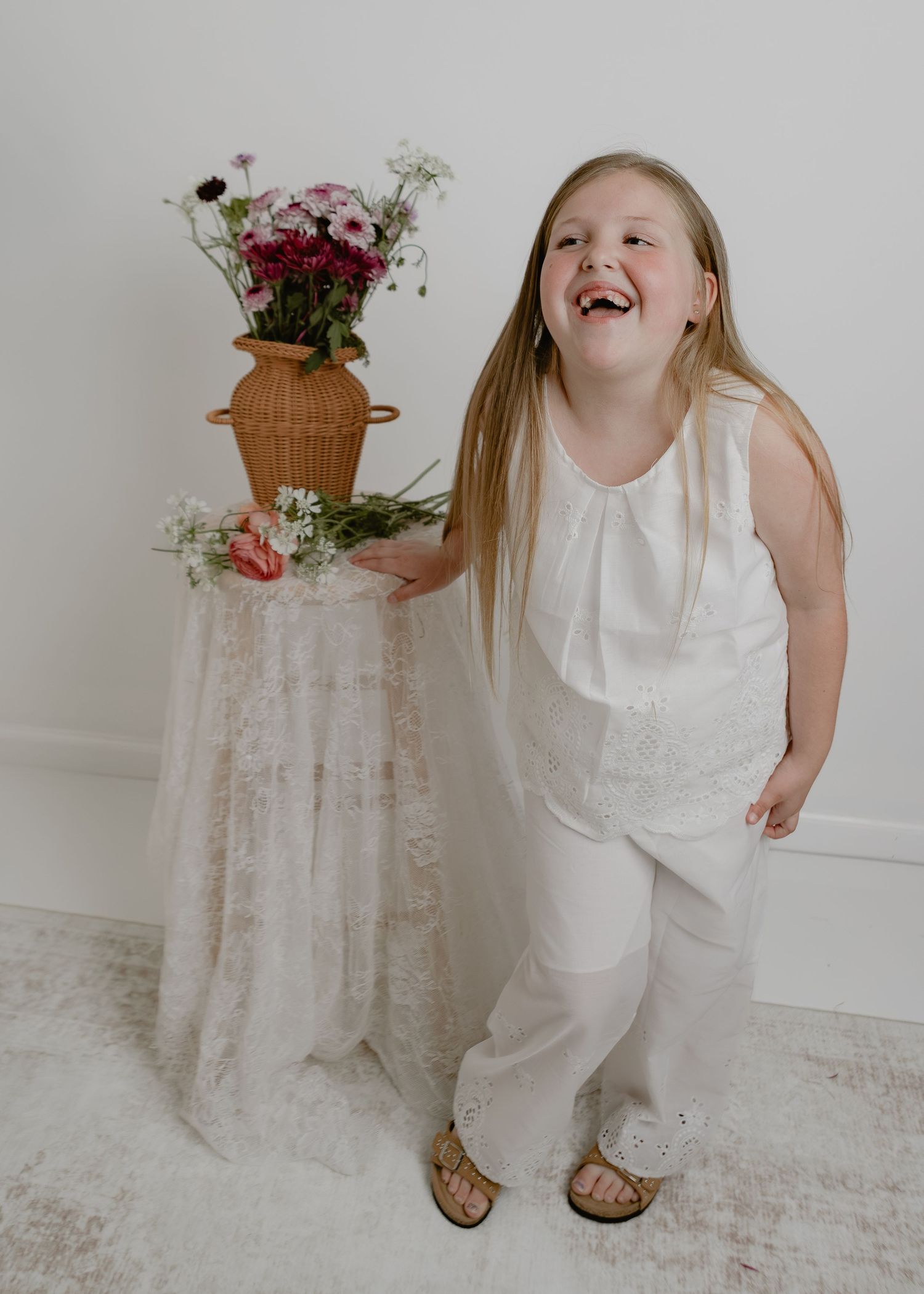 Joyful young girl in white outfit laughing beside a floral arrangement in a bright studio.