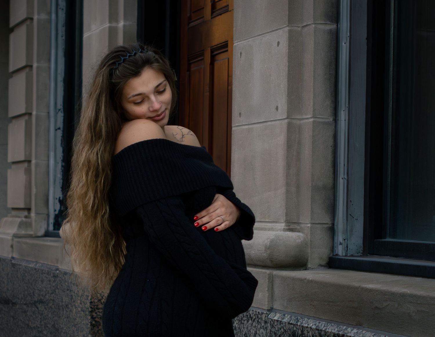 Young woman with long curly hair in a black off-shoulder top leaning against a stone building.