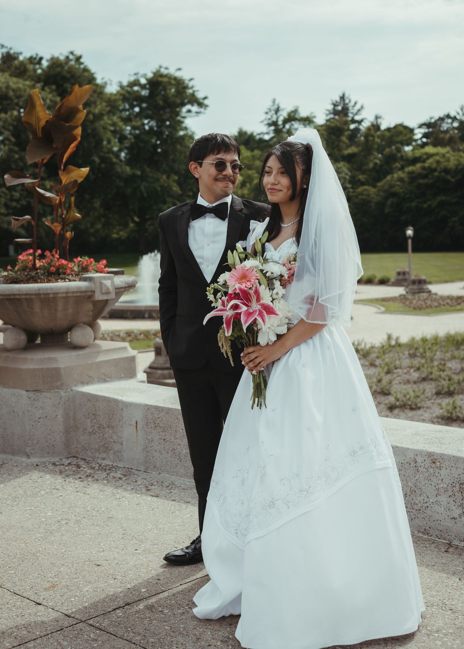 Bride and groom posing outdoors, bride in white gown with veil holding pink lily bouquet, groom in black tuxedo.